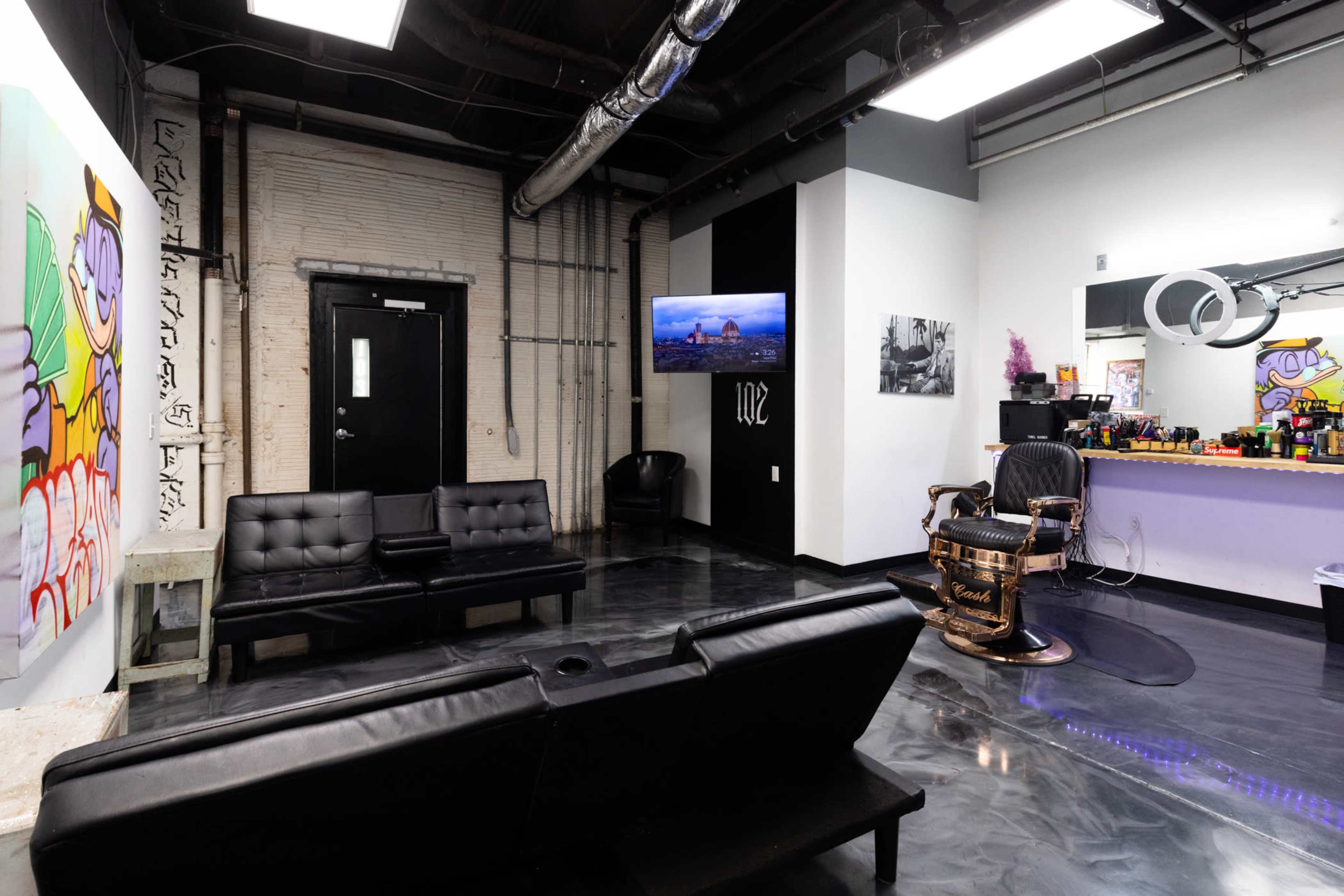 The image shows a modern barber shop interior featuring black leather seating, a vibrant wall art piece, a television mounted on the wall, and a barber chair in front of a brightly colored reception area.