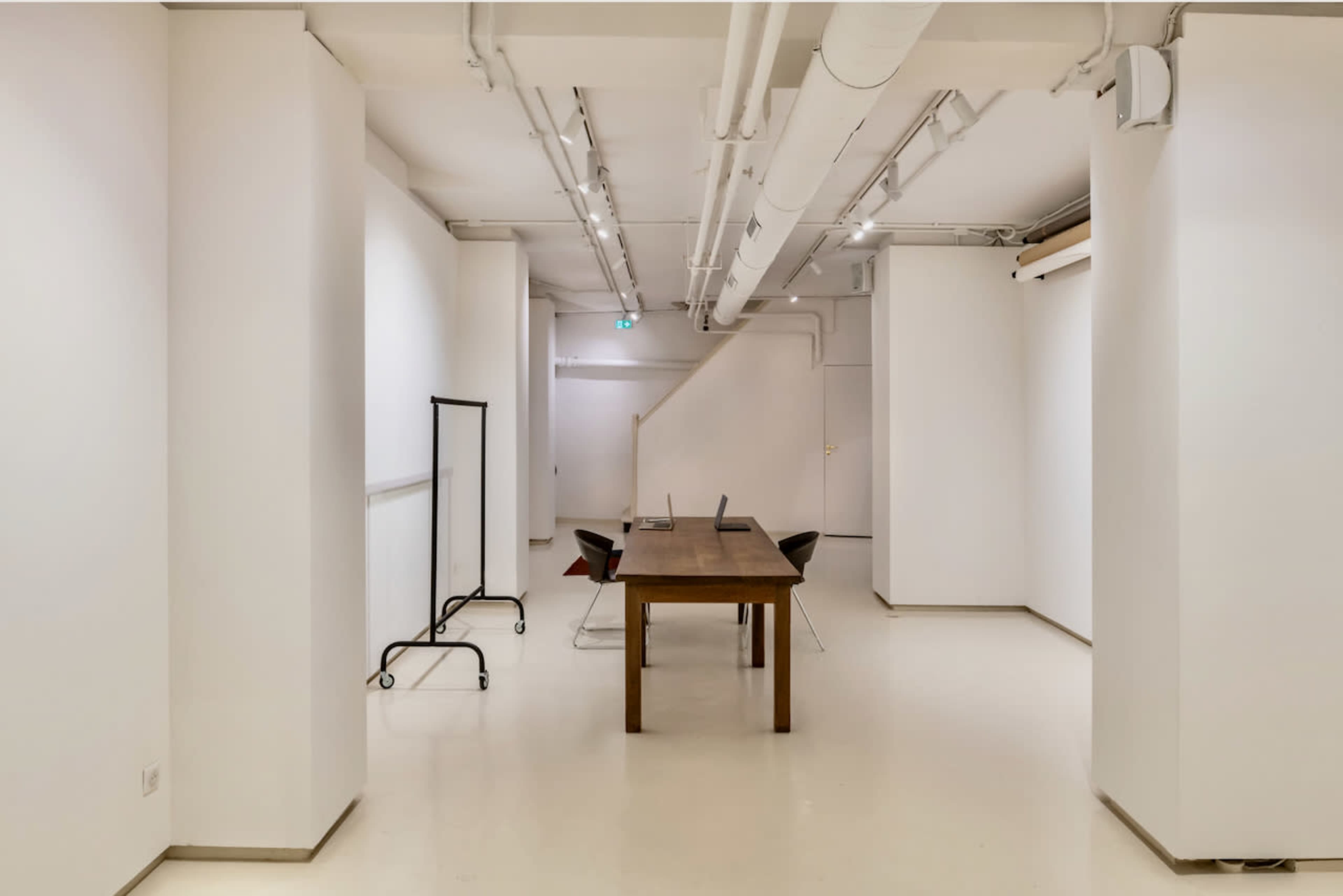 A spacious, minimalistic room features a long wooden table surrounded by chairs, with white walls and exposed pipes.
