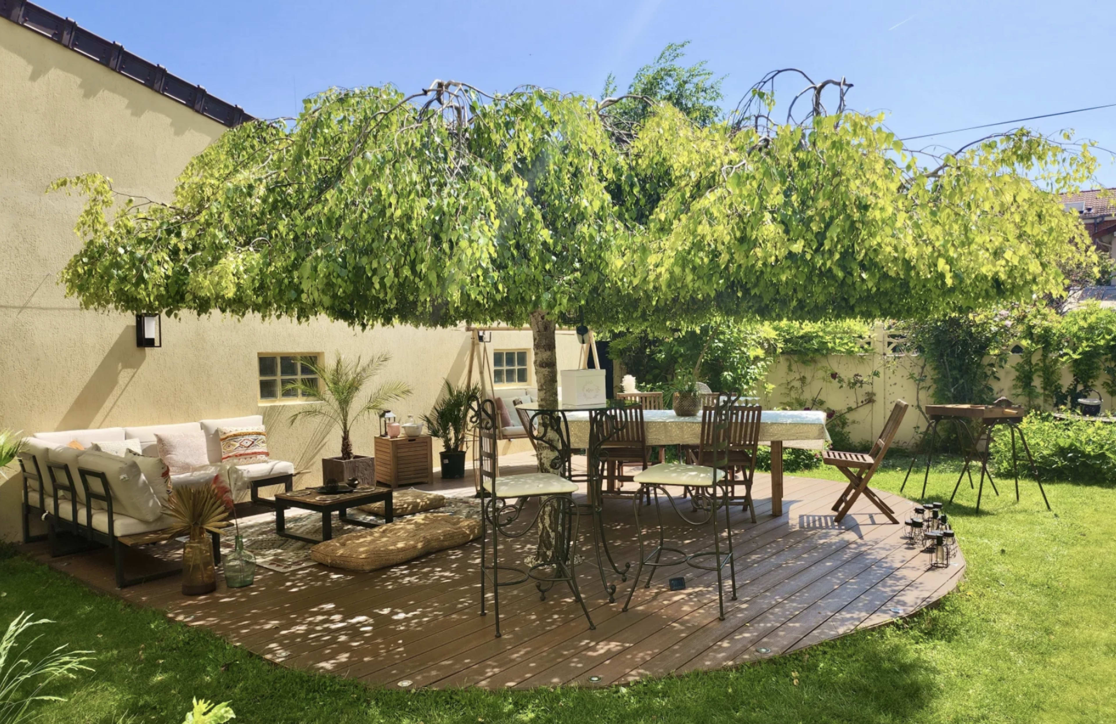 A wooden patio with a round table and chairs is shaded by a large tree, surrounded by green grass and garden decor.