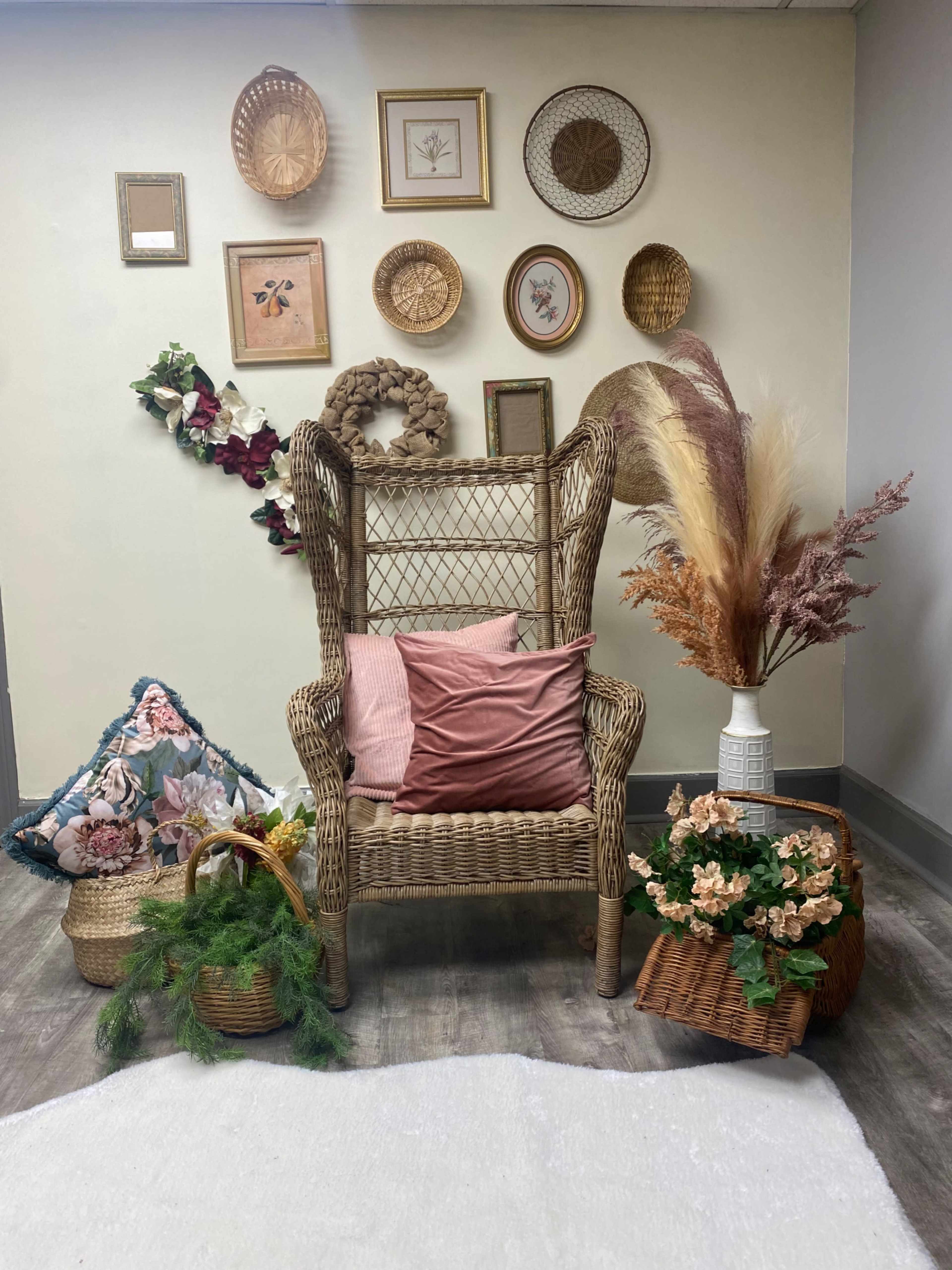 A wicker chair is surrounded by decorative baskets, floral arrangements, and framed pictures against a wall.