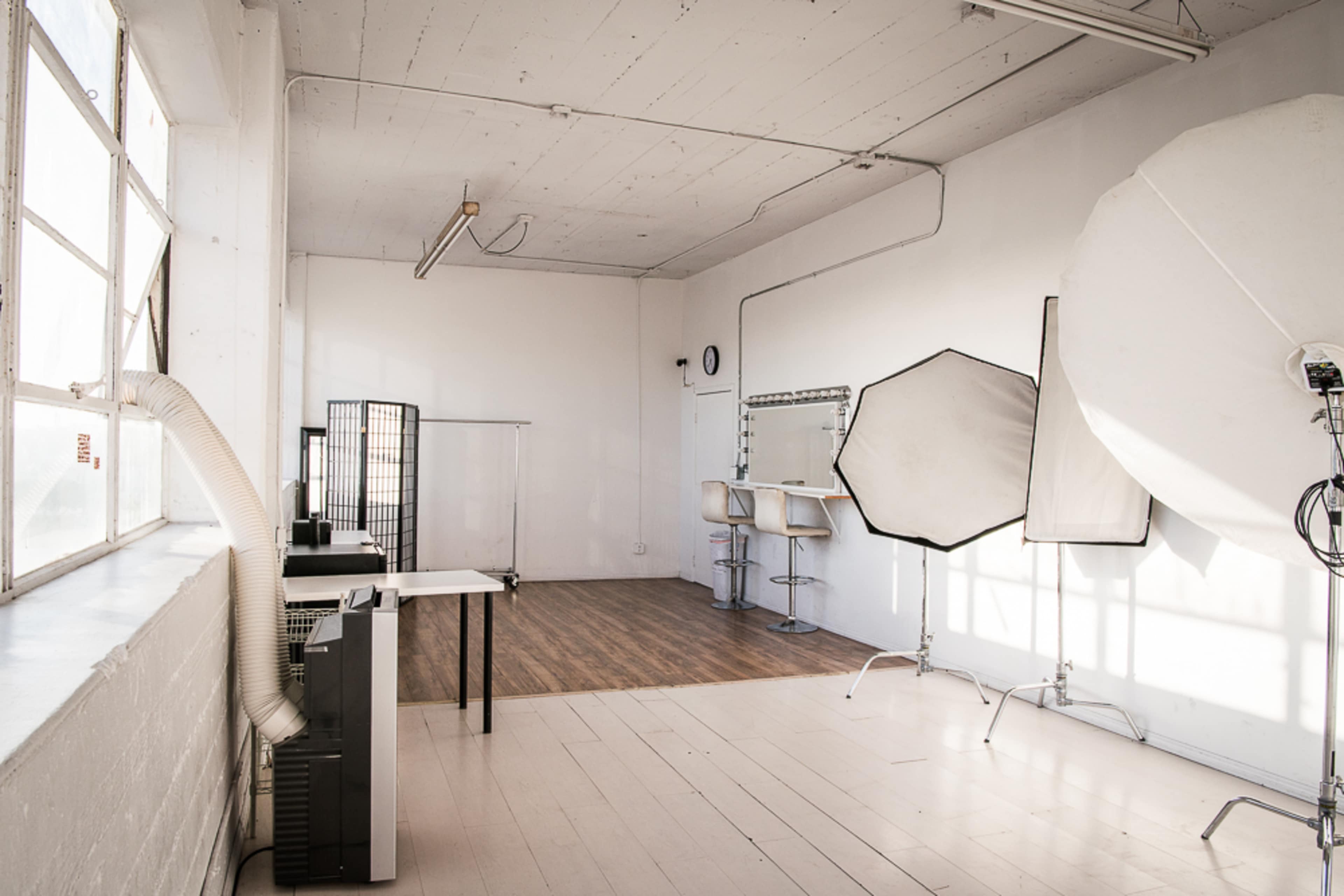 The image shows a photography studio with large windows, a light-colored floor, and various equipment including a backdrop, a table, and lighting modifiers.