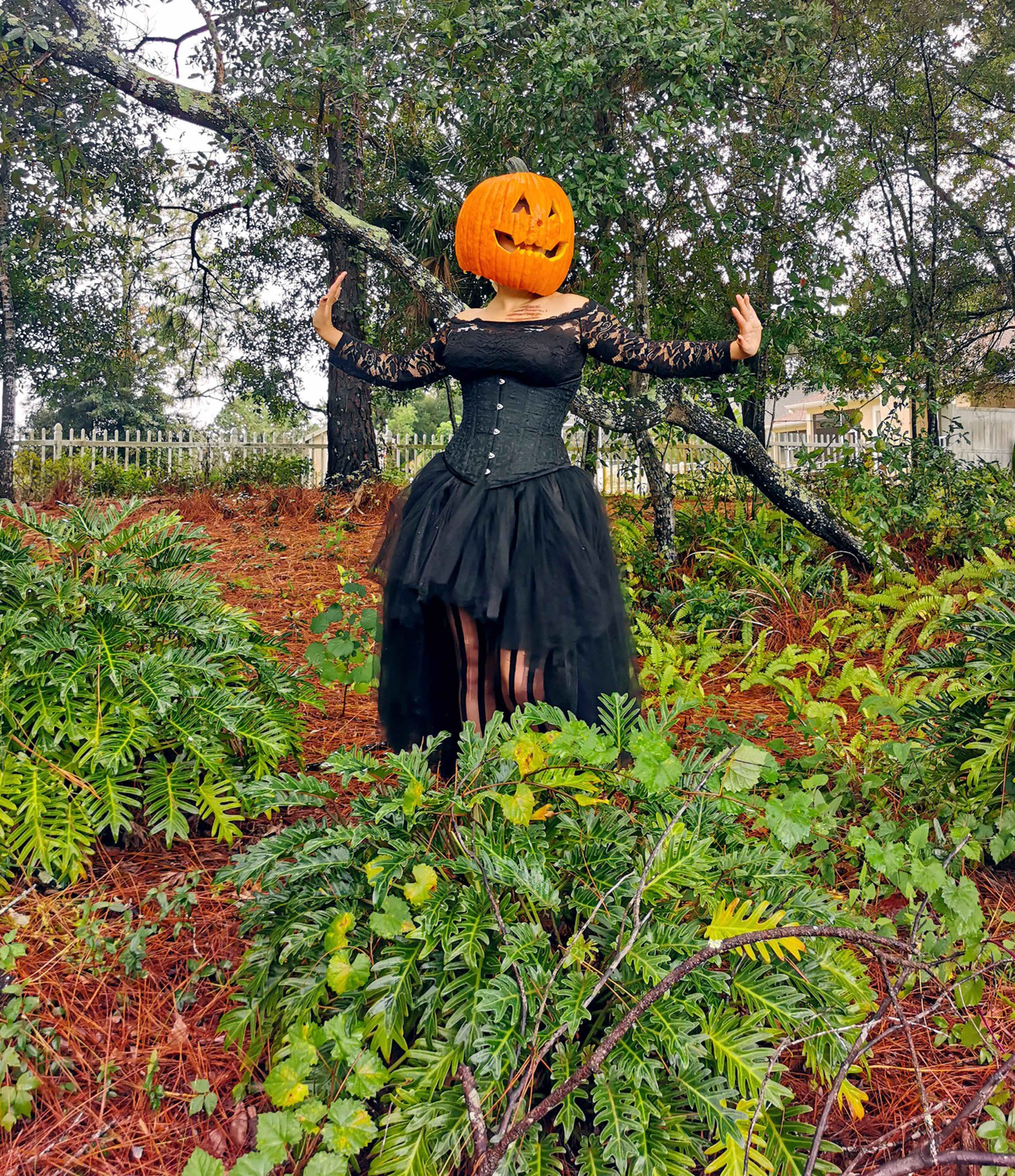 A person wearing a black dress and a pumpkin head stands among green foliage in a wooded area.