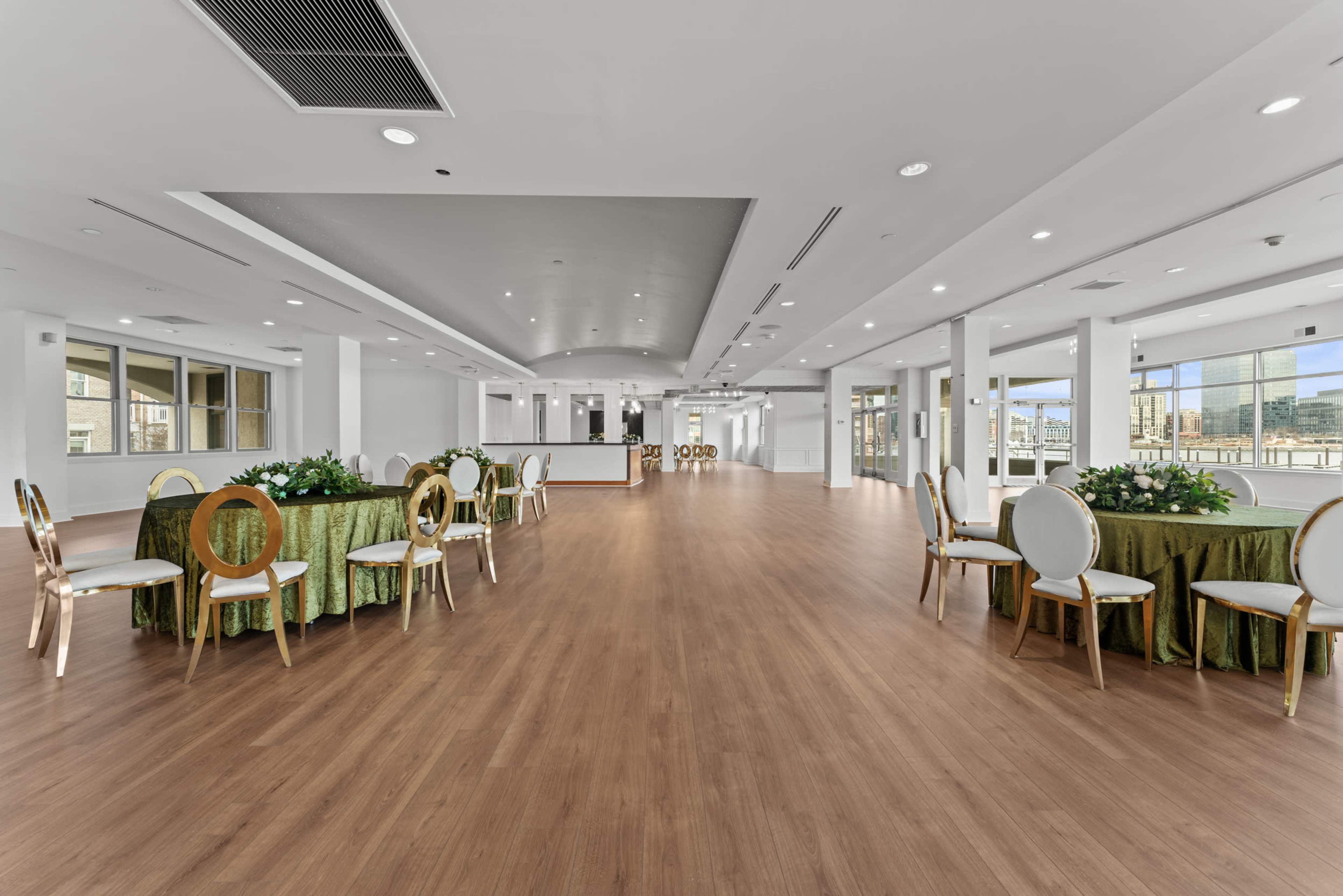 The image shows an empty, well-lit event space featuring several round tables with green tablecloths and elegant chairs arranged around them.