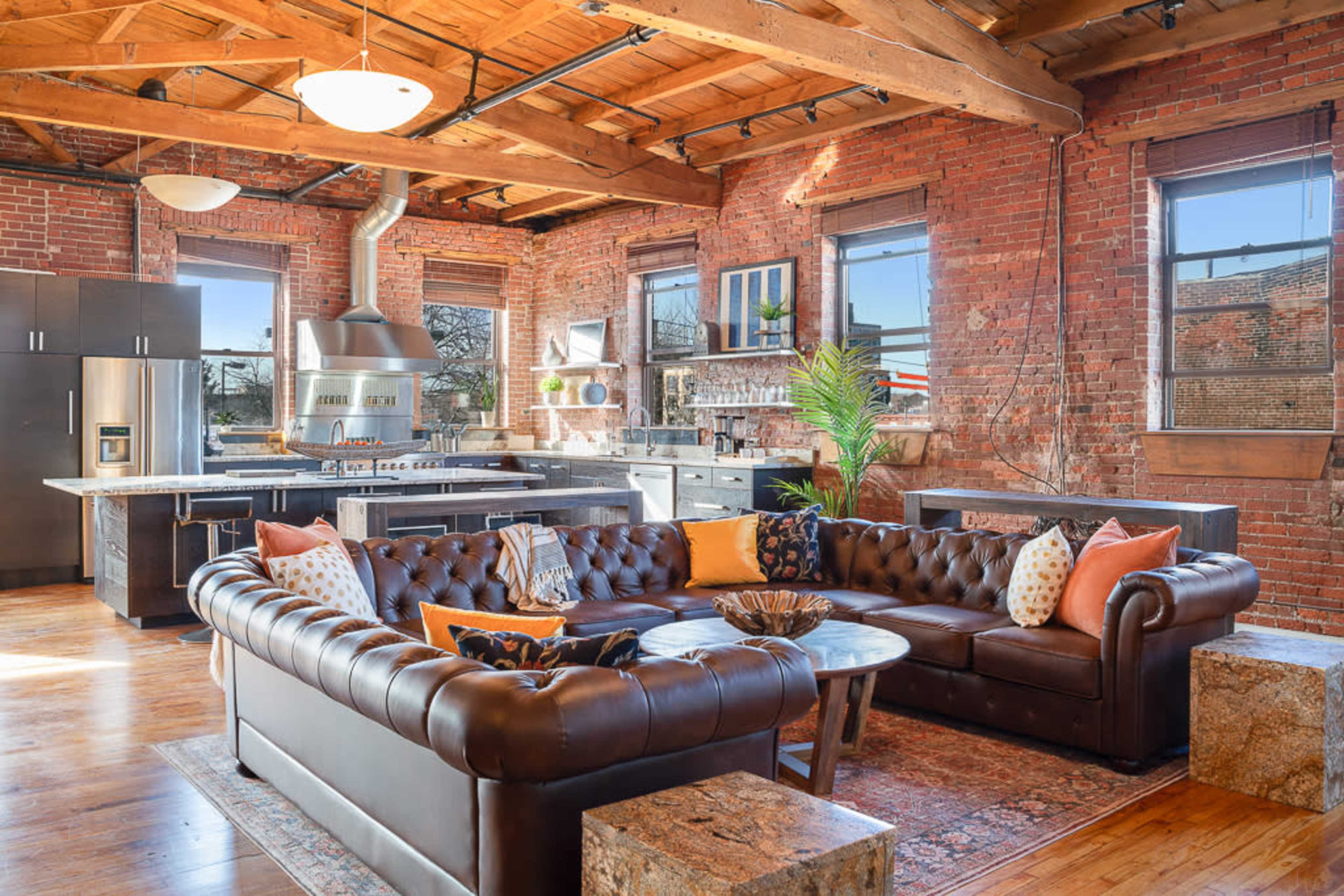 A spacious kitchen and living area features a large brown leather sectional sofa, a round coffee table, and exposed brick walls, with modern appliances visible in the background.