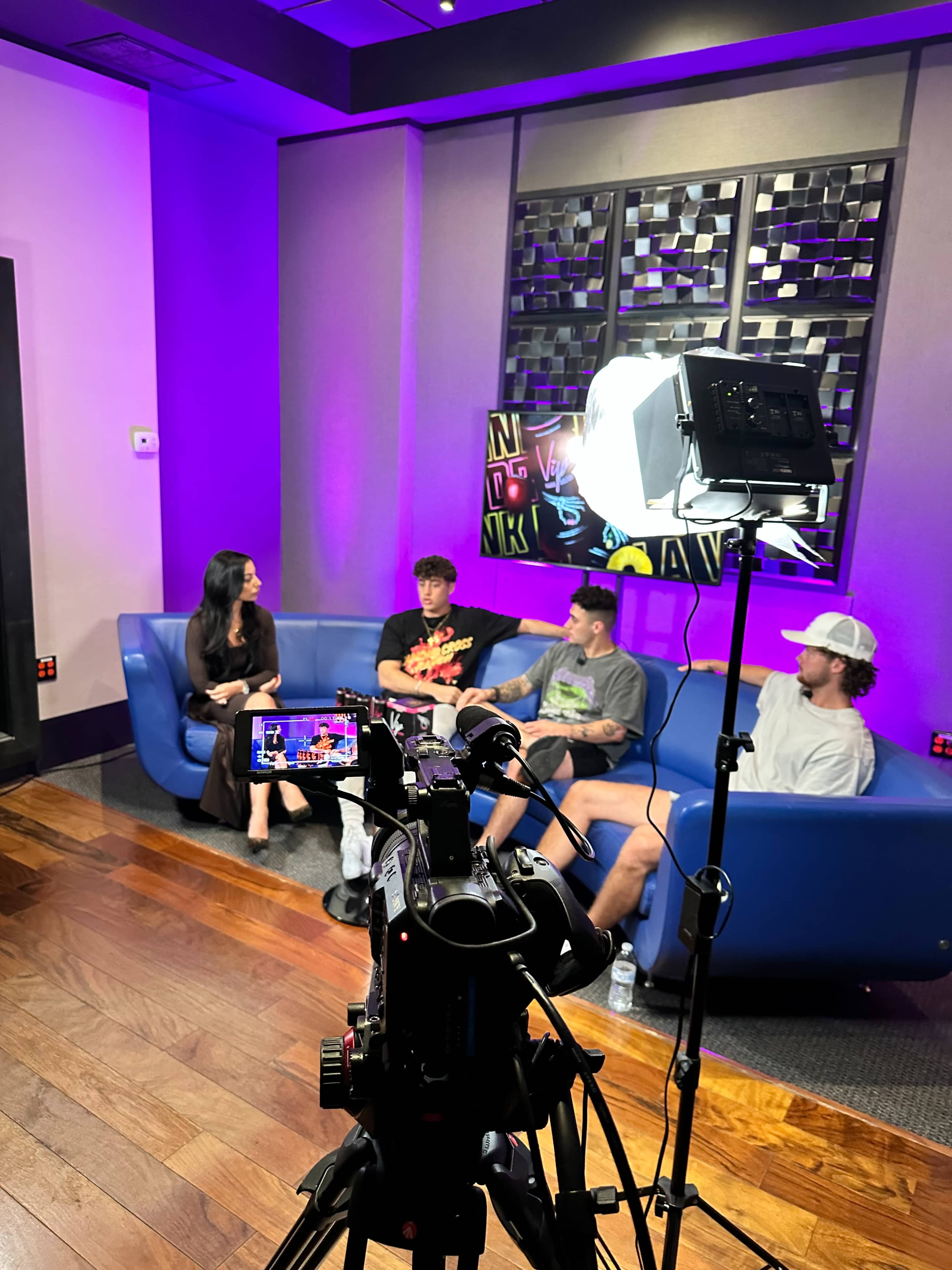 A group of four individuals sits on a blue couch in a well-lit room, with a camera set up in front of them for recording.