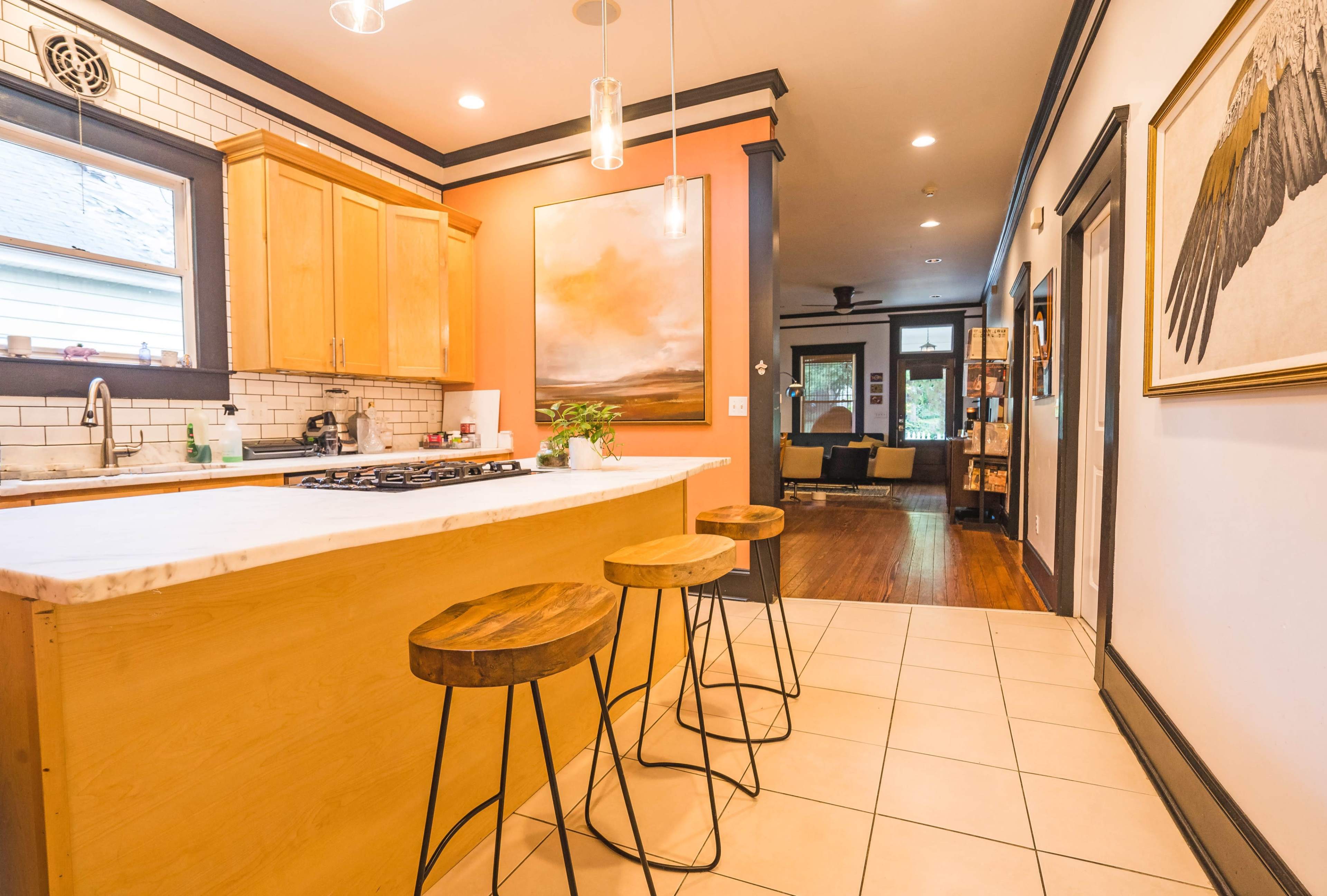A contemporary kitchen with wooden cabinetry, a marble countertop, and three metal bar stools, leading into a living area with a sofa and artwork on the walls.