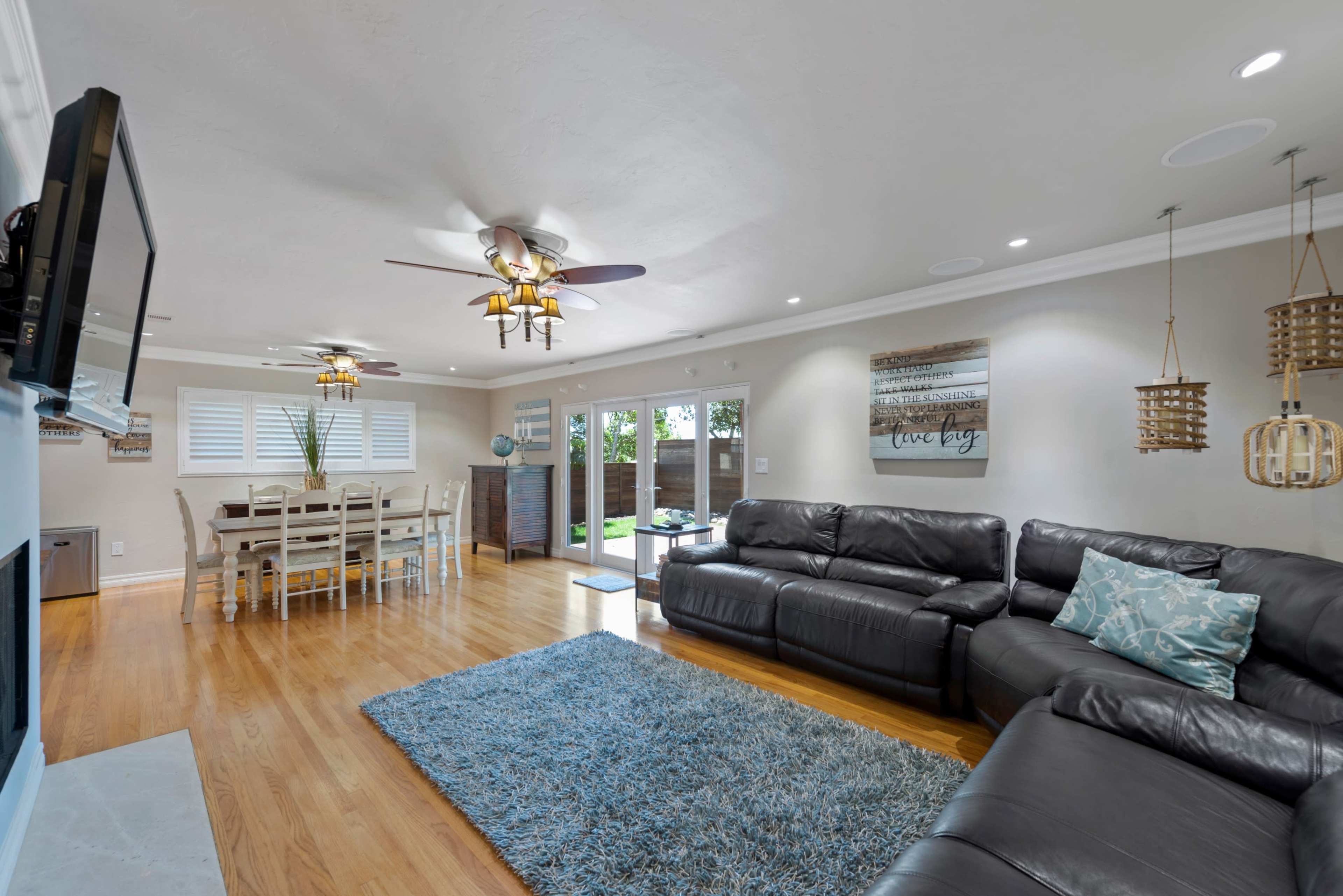 The image shows a spacious living room with a large black sectional sofa, a wooden dining table, and access to a backyard through sliding glass doors.