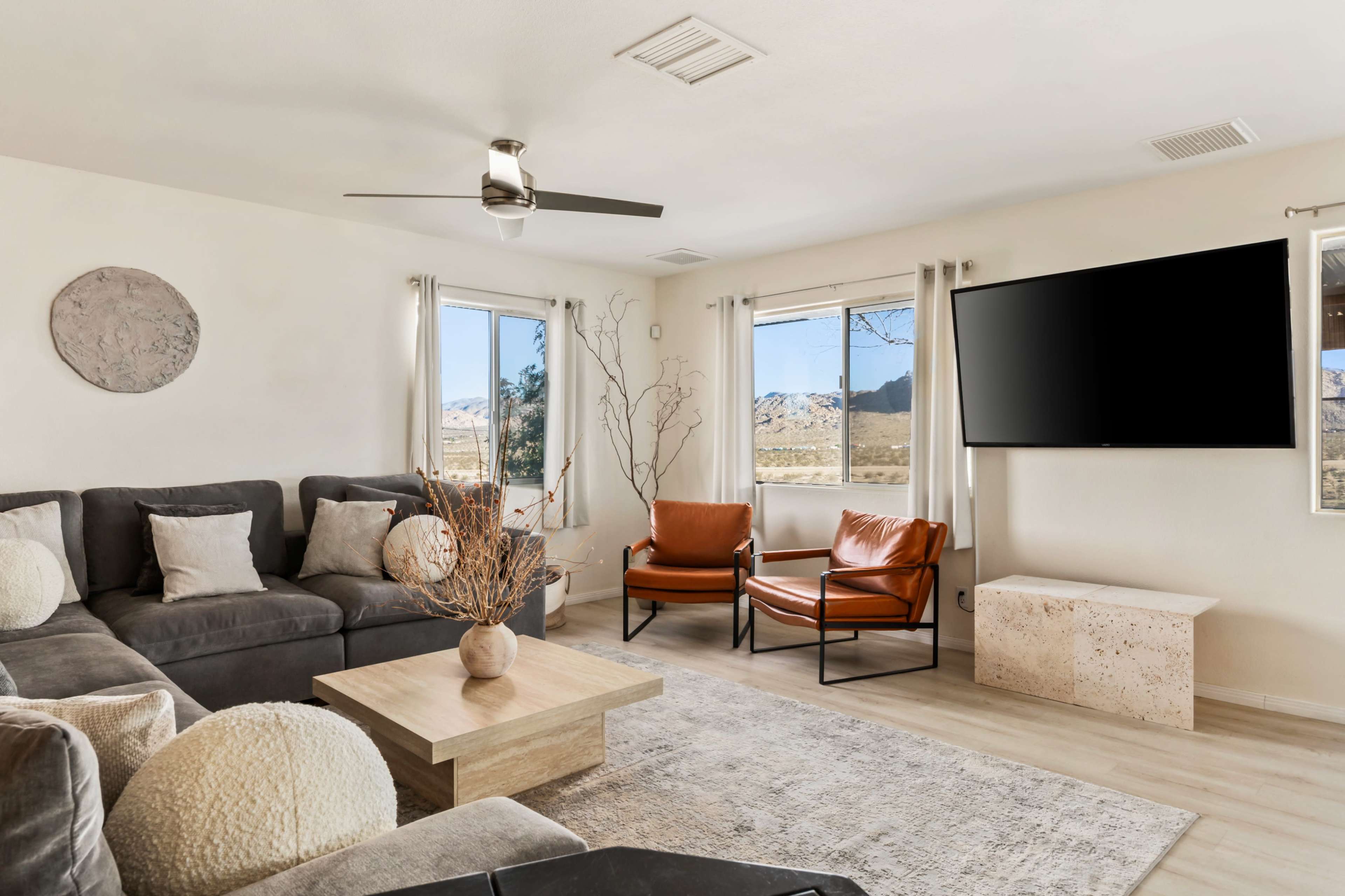 The image shows a modern living room with a sectional sofa, two leather armchairs, a coffee table, and a wall-mounted television, surrounded by large windows that offer a view of the outdoors.