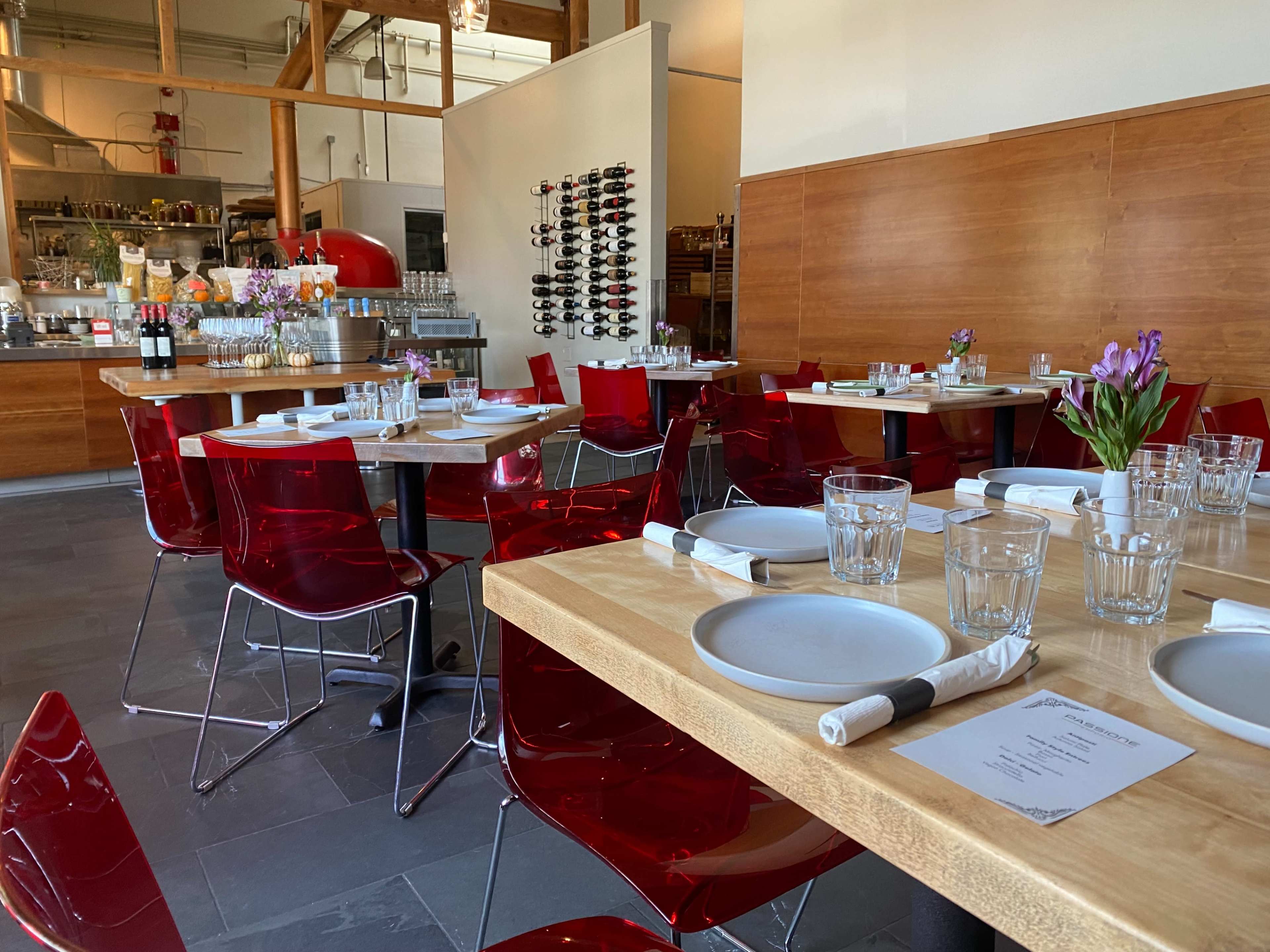A modern restaurant interior with wooden tables, red chairs, and a wine rack in the background.