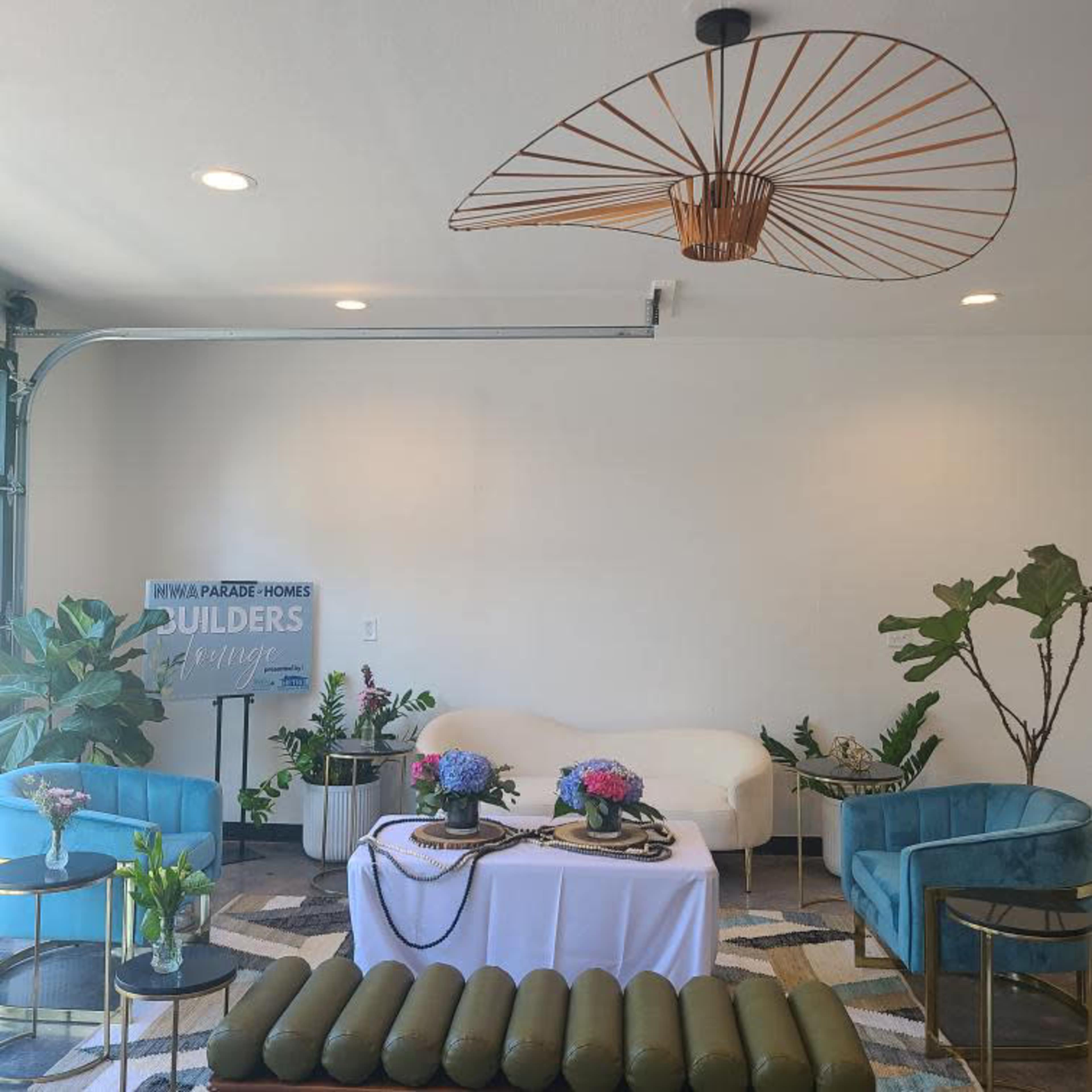 The image shows a stylish interior space featuring a white sofa, two blue armchairs, a decorative ceiling fixture, potted plants, and a table with floral arrangements.