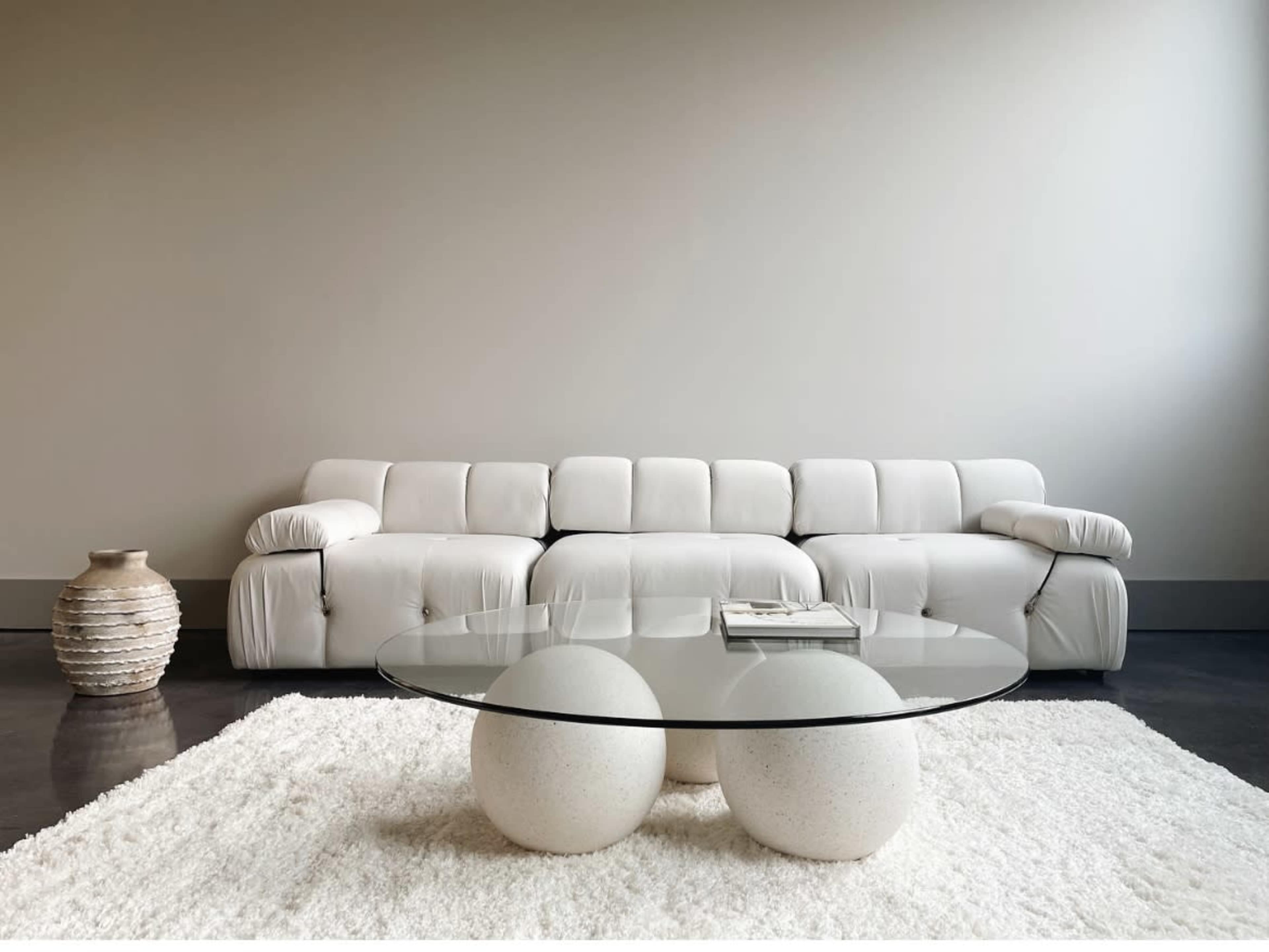 A modern living room features a white sectional sofa, a round glass coffee table with two stone bases, and a textured area rug.