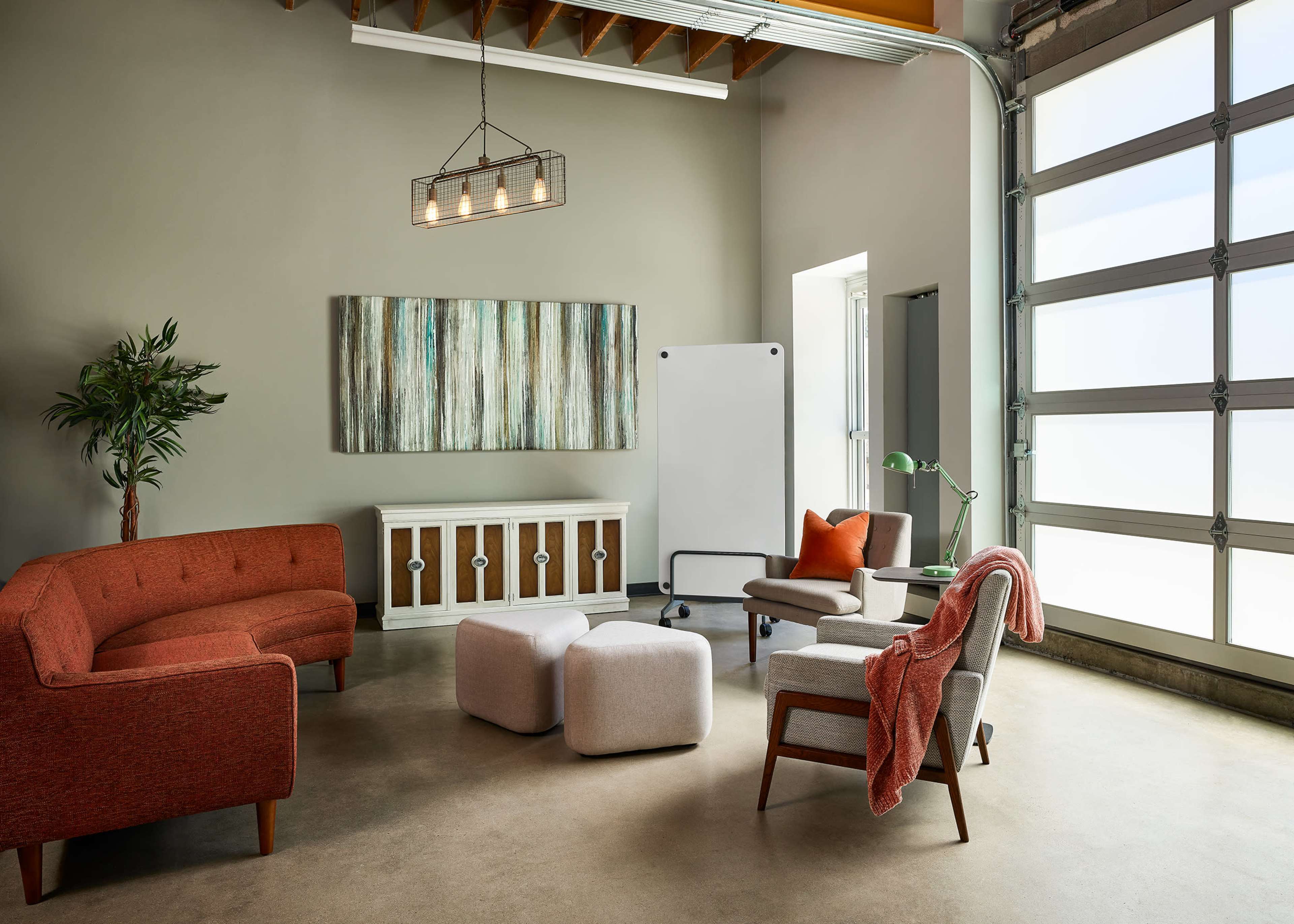 A modern office space features a seating area with a brown sofa, several ottomans, a whiteboard, and large windows allowing natural light.