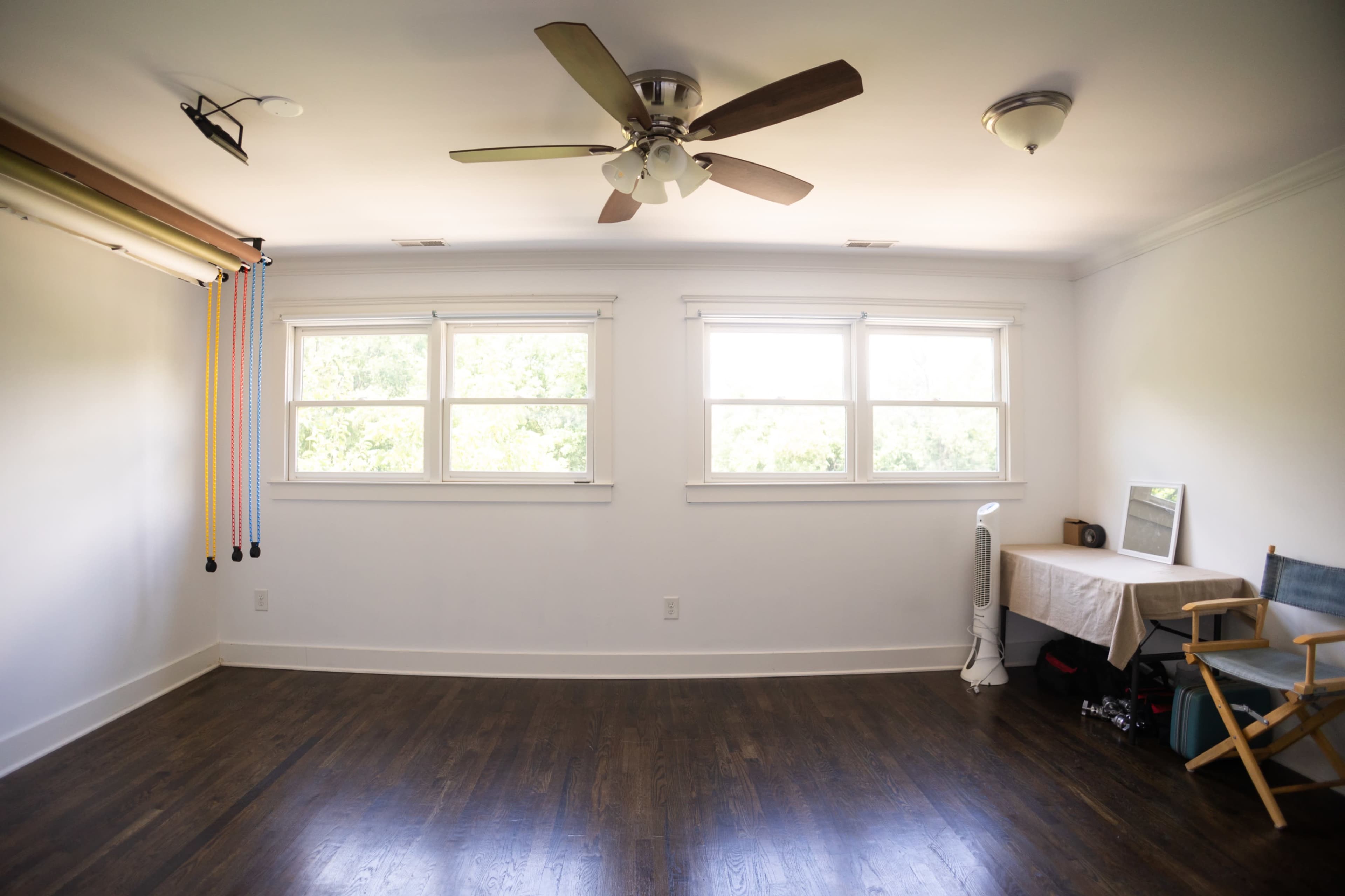 The image shows a simplified, well-lit room with a ceiling fan, two windows, a table, a chair, and exercise equipment along one wall.