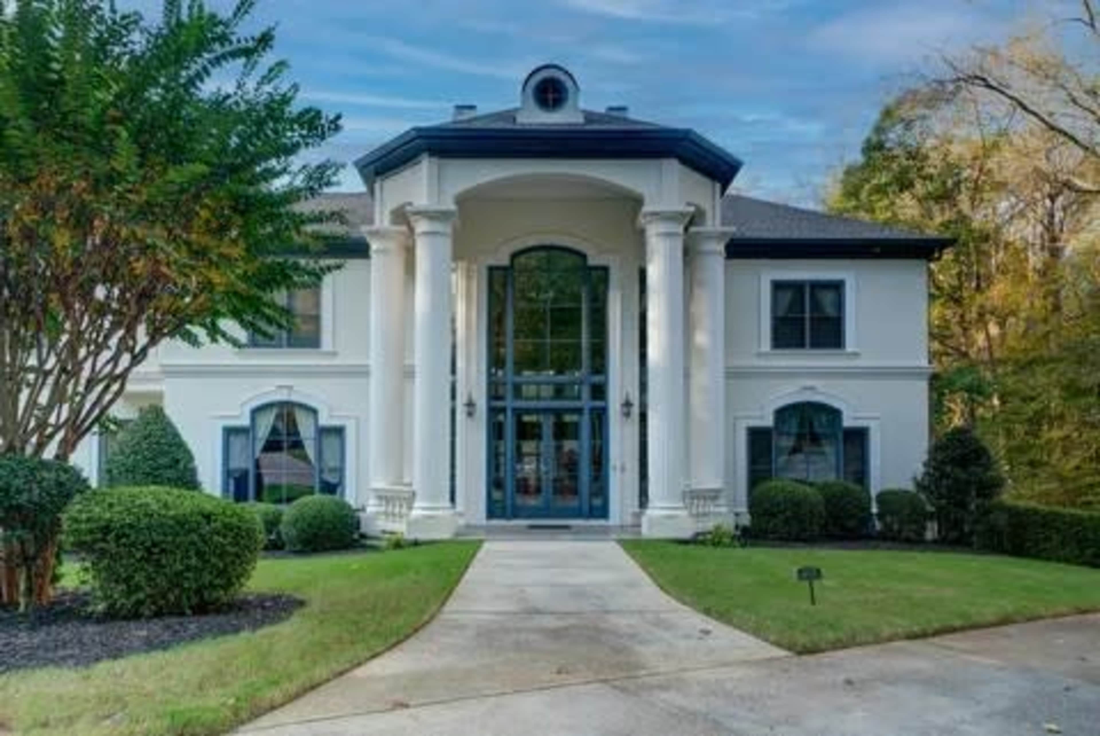 The image shows a large, modern white house with two prominent columns at the entrance, surrounded by neatly trimmed bushes and a well-maintained lawn.