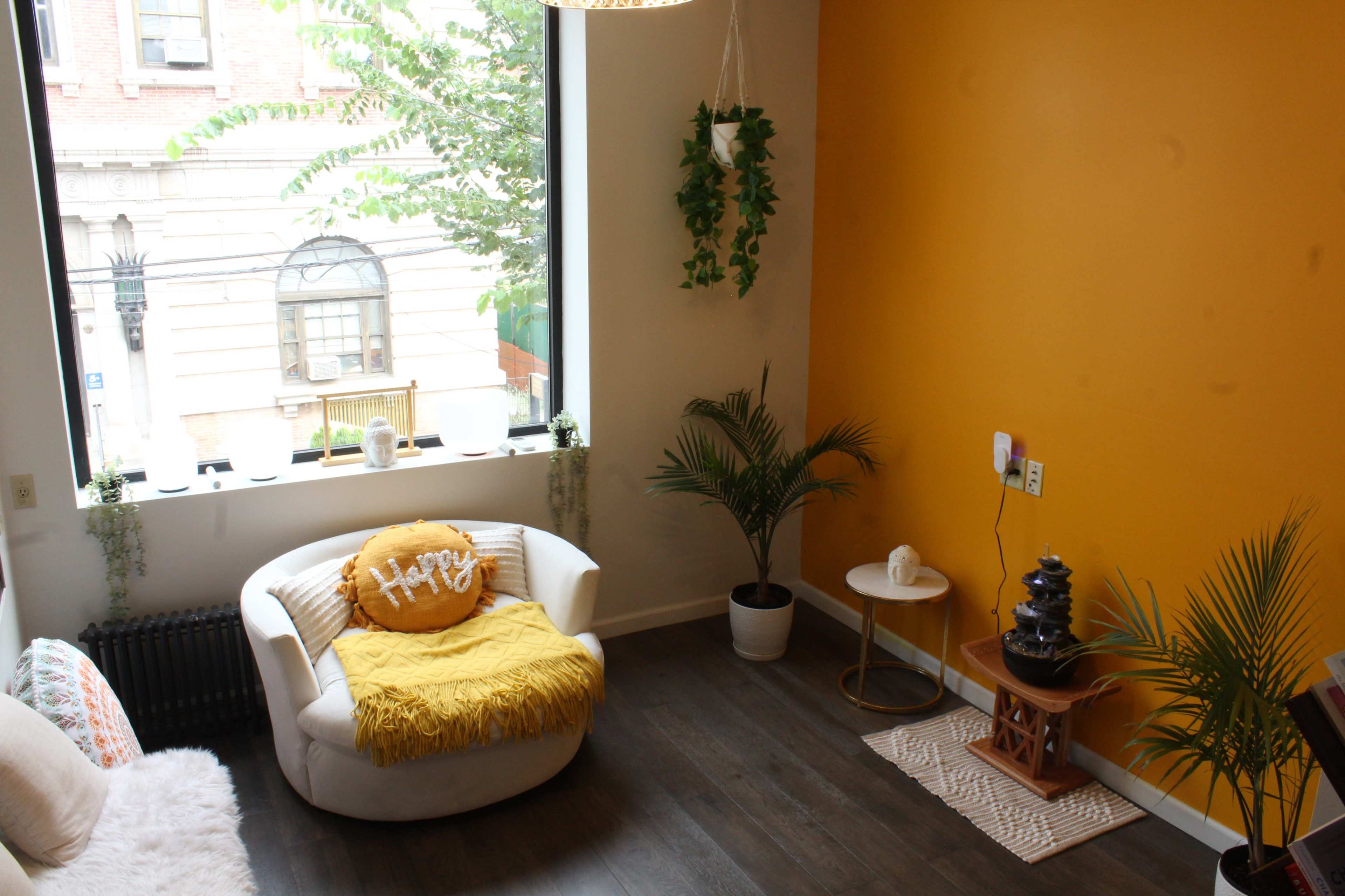 The image shows a small, bright room with a white armchair, a decorative pillow, a plant in a pot, a small side table, and a water feature against a warm yellow wall.