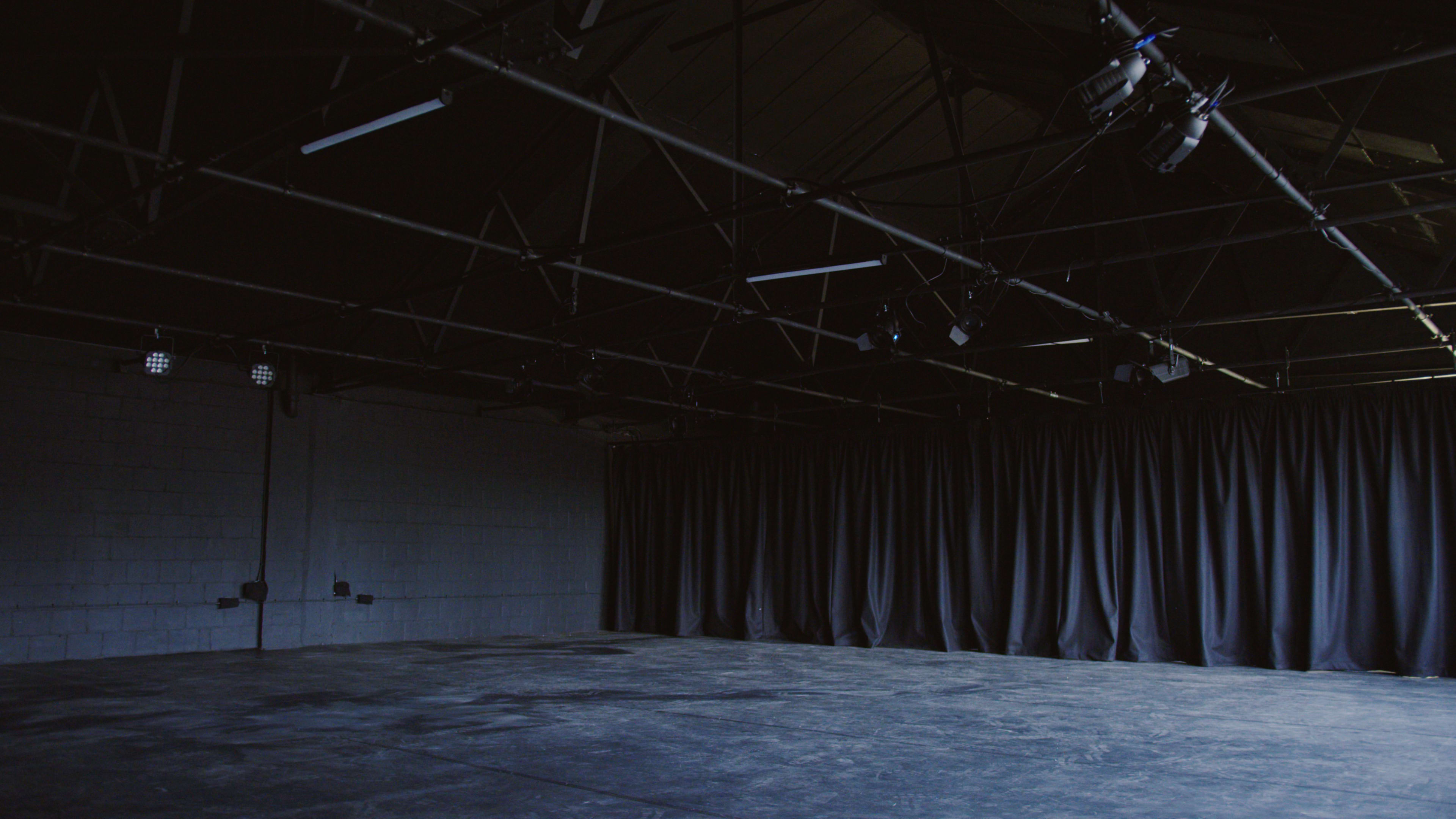 An empty stage area with a dark floor and black curtains behind, under a ceiling with visible lighting fixtures.