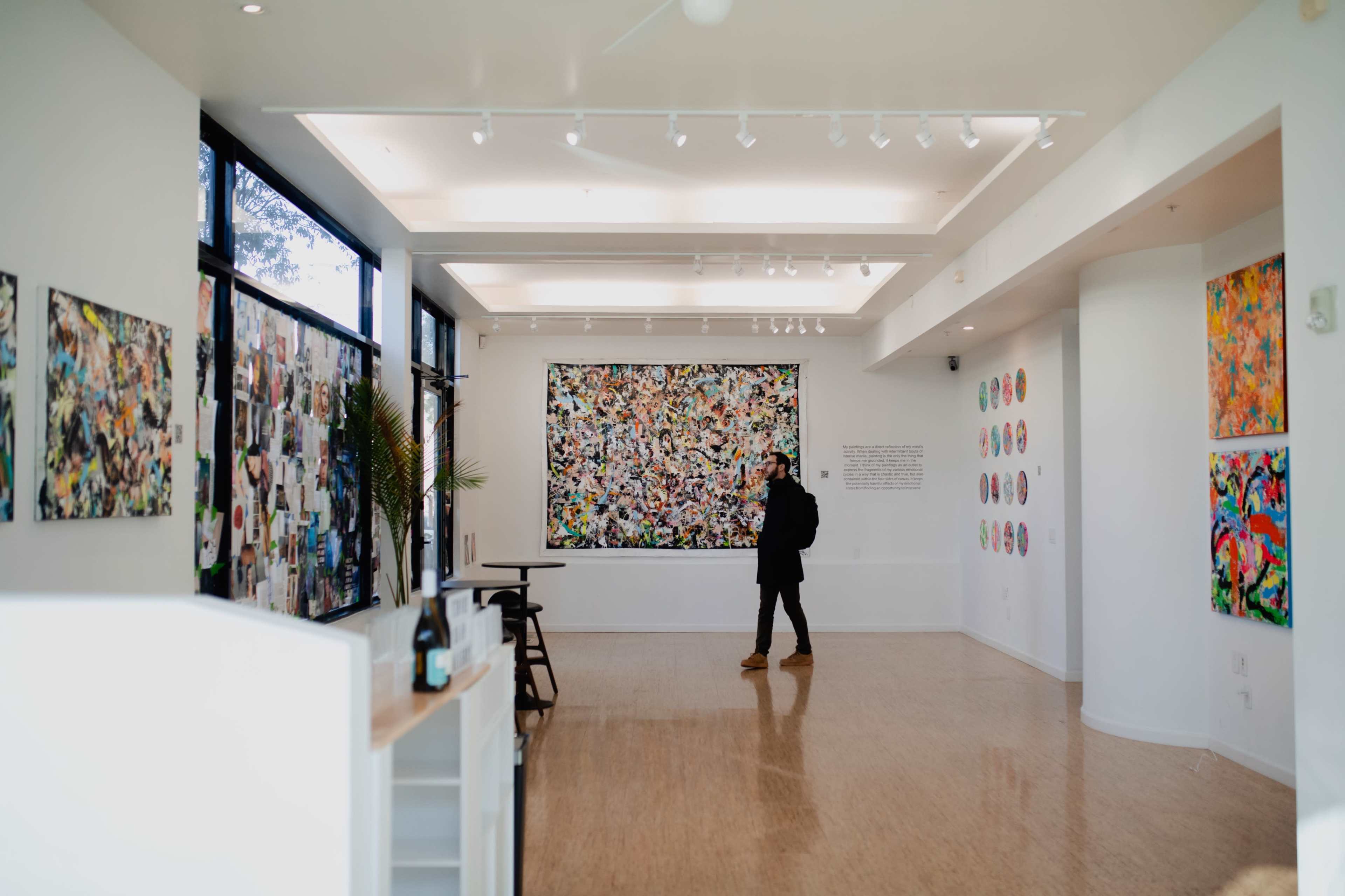 A person stands in an art gallery, observing a large colorful artwork on the wall, surrounded by other art pieces and photographs displayed on the walls.
