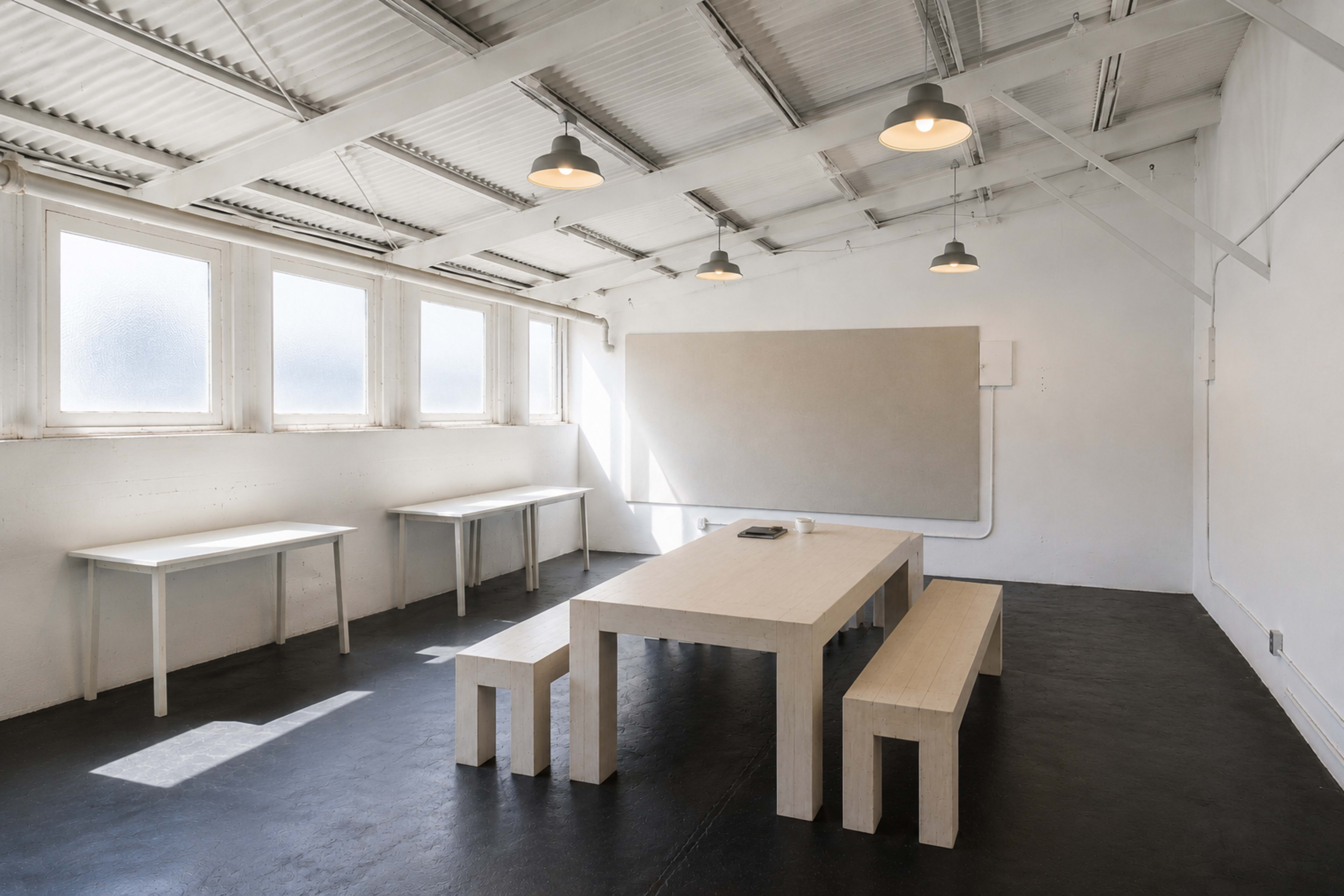The image shows a minimalist room with white walls, two tables, benches, and a large whiteboard at one end.