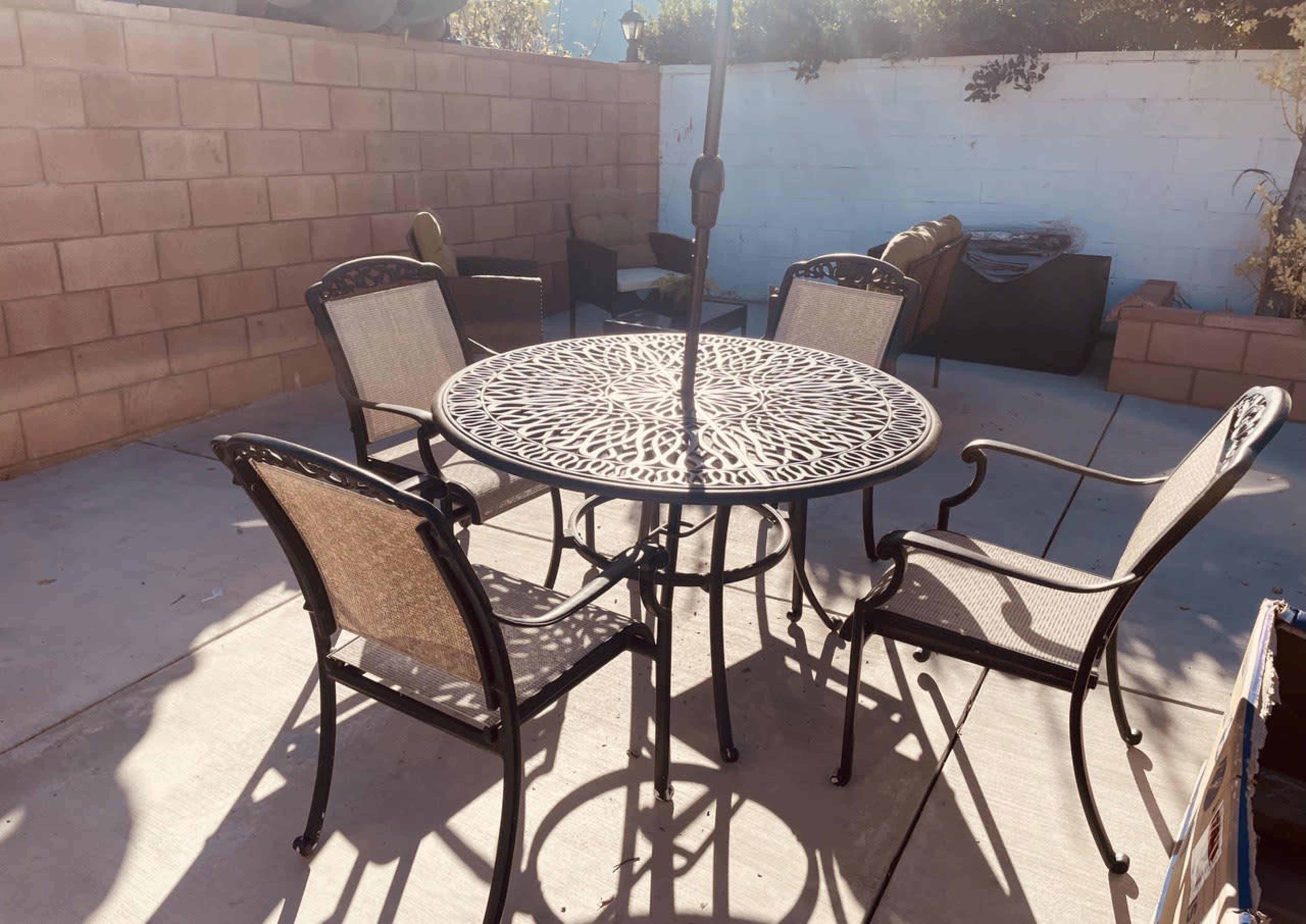 A round metal table with ornate patterns is surrounded by four mesh chairs on a concrete patio, with a wall in the background.