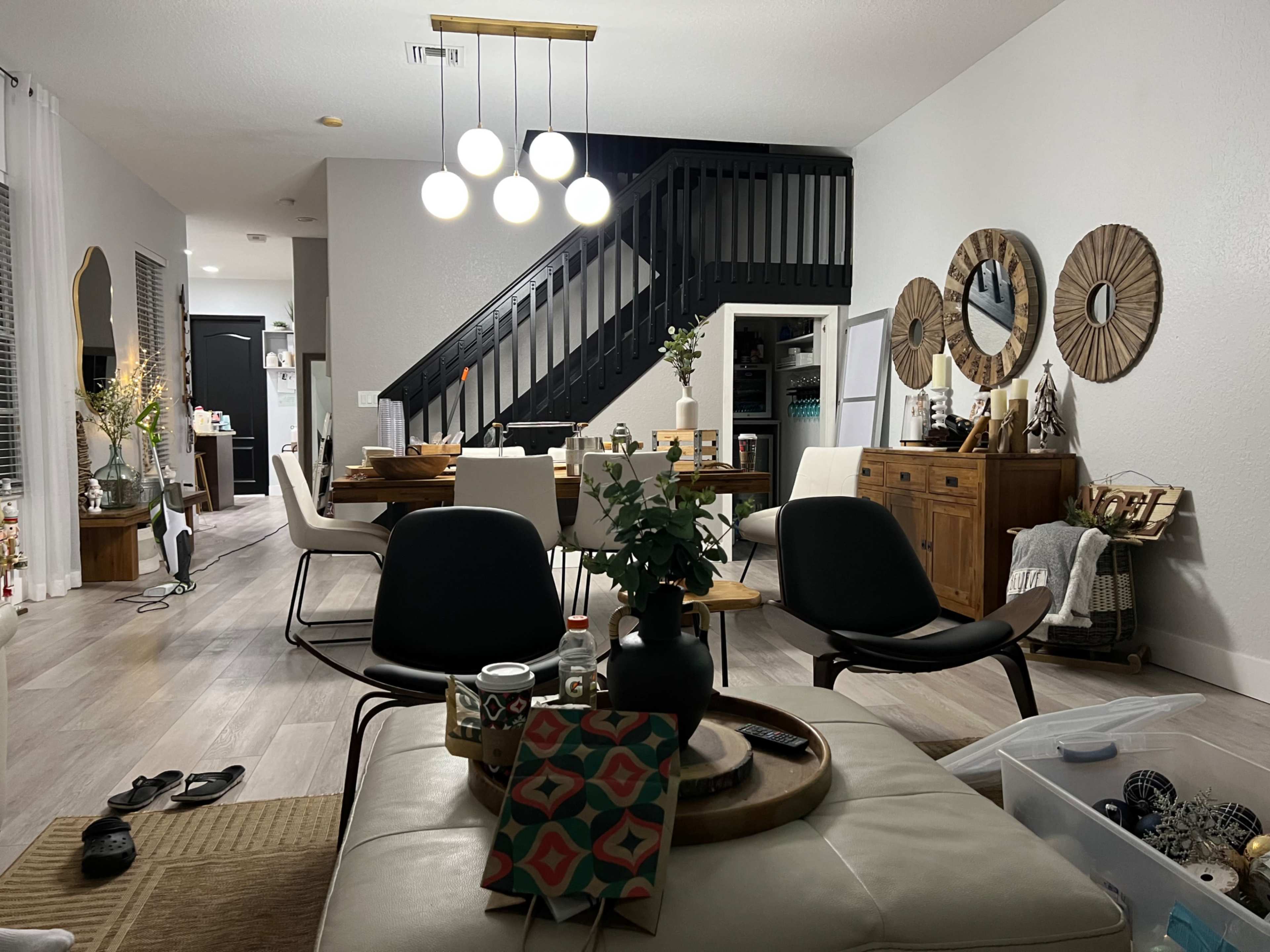 A modern living and dining area featuring a staircase, a dining table set for six, decorative mirrors on the walls, and various household items arranged on furniture.