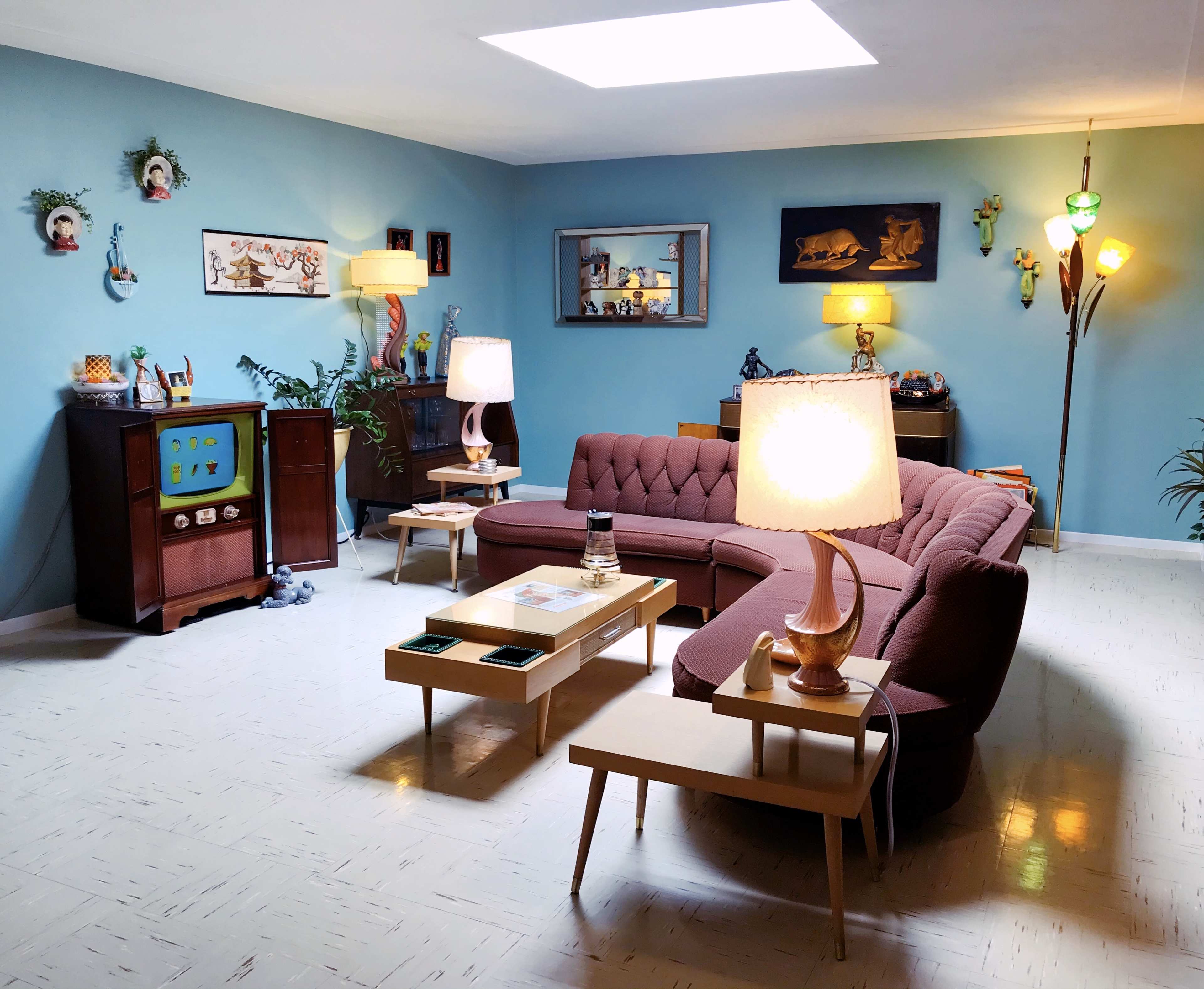 A brightly lit retro living room featuring a pink sectional sofa, a vintage television set, wooden side tables, and various decorative items on the walls.
