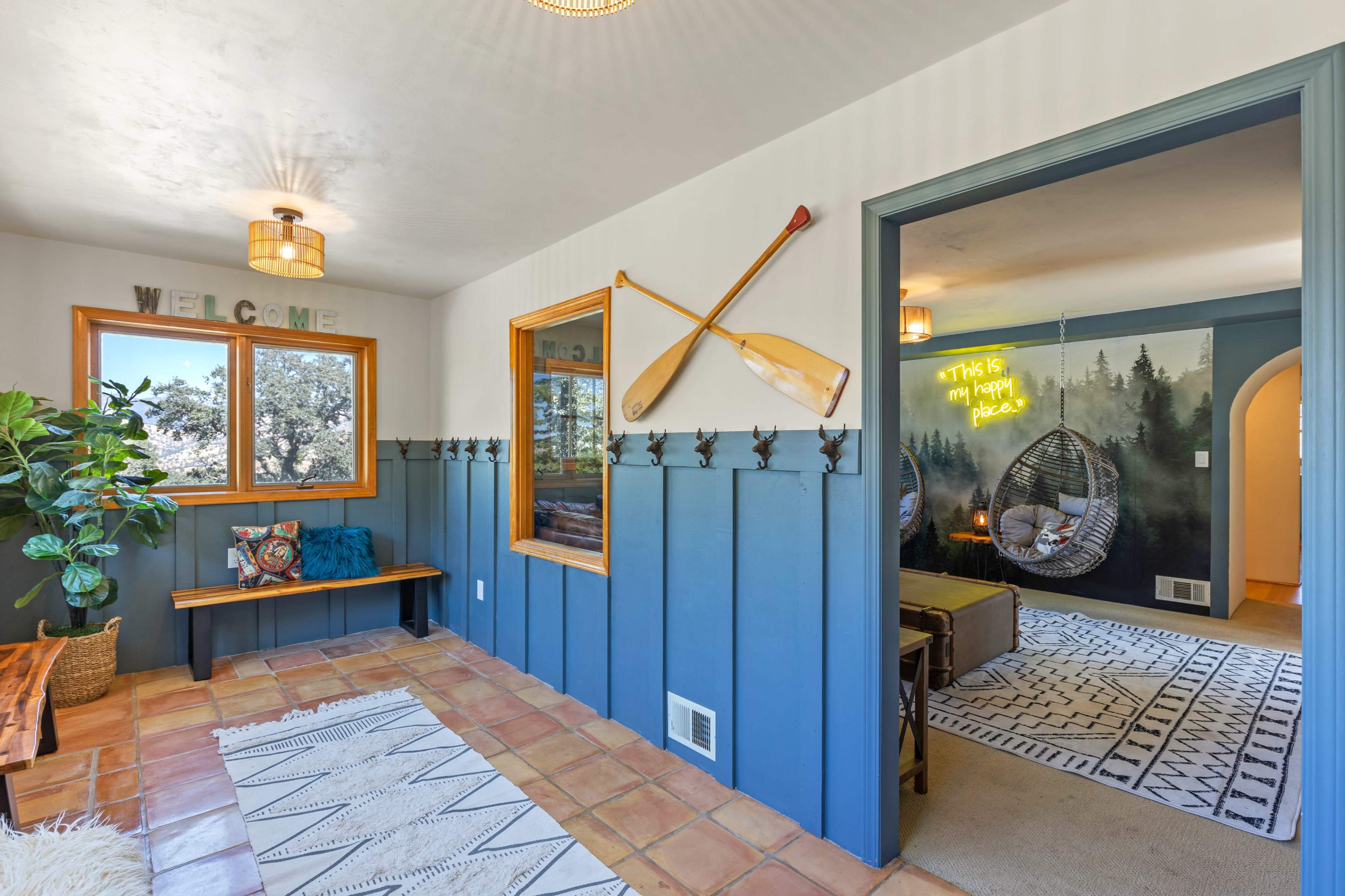 An entryway with a wooden bench, coat hooks on the wall, and a bright room visible through an open doorway featuring hanging chairs and decorative wall art.