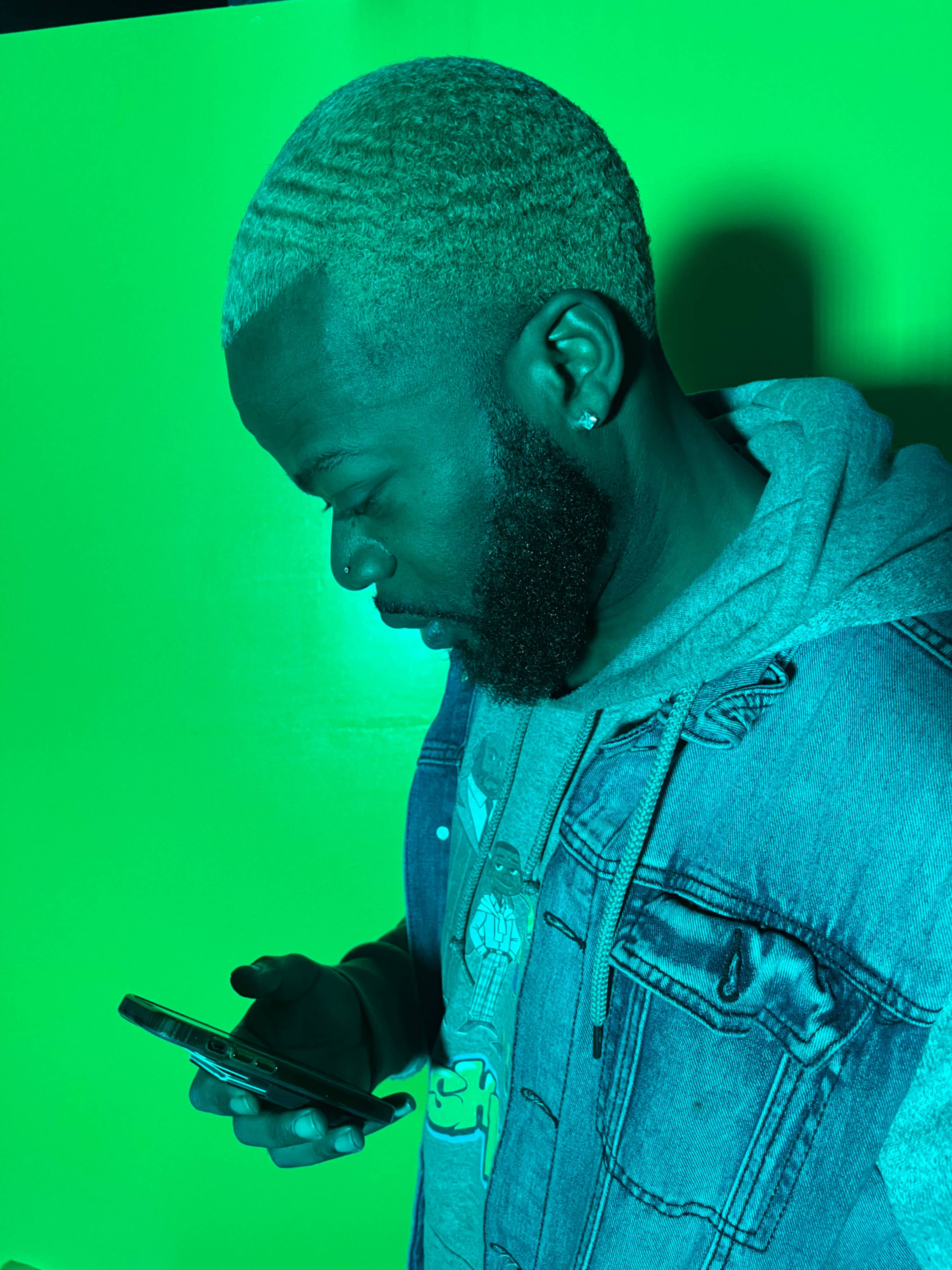 A man with blonde, wavy hair and a beard is looking at his smartphone against a green background.