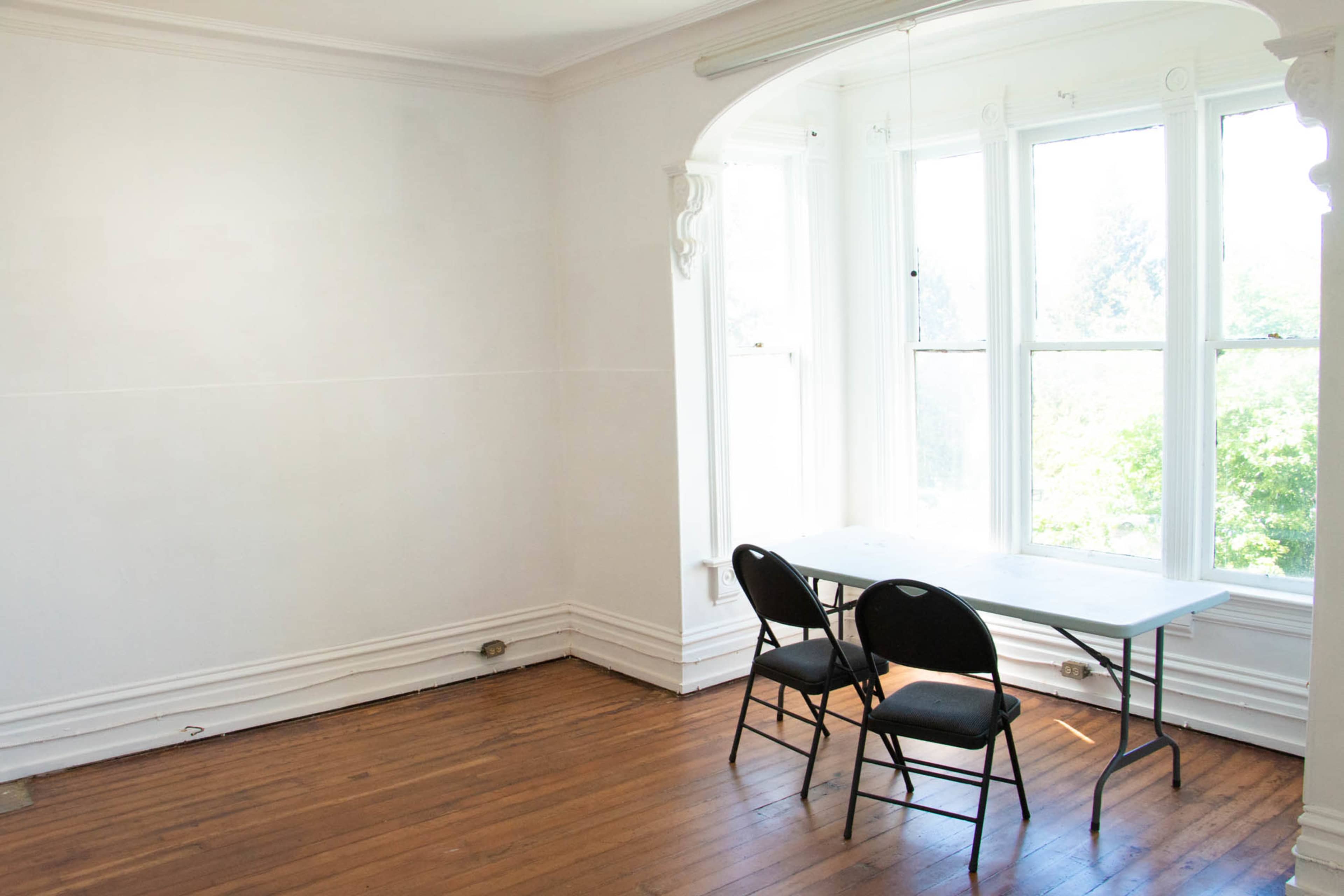 A sparsely furnished room features a table and two black chairs near a large window with a view of greenery outside.