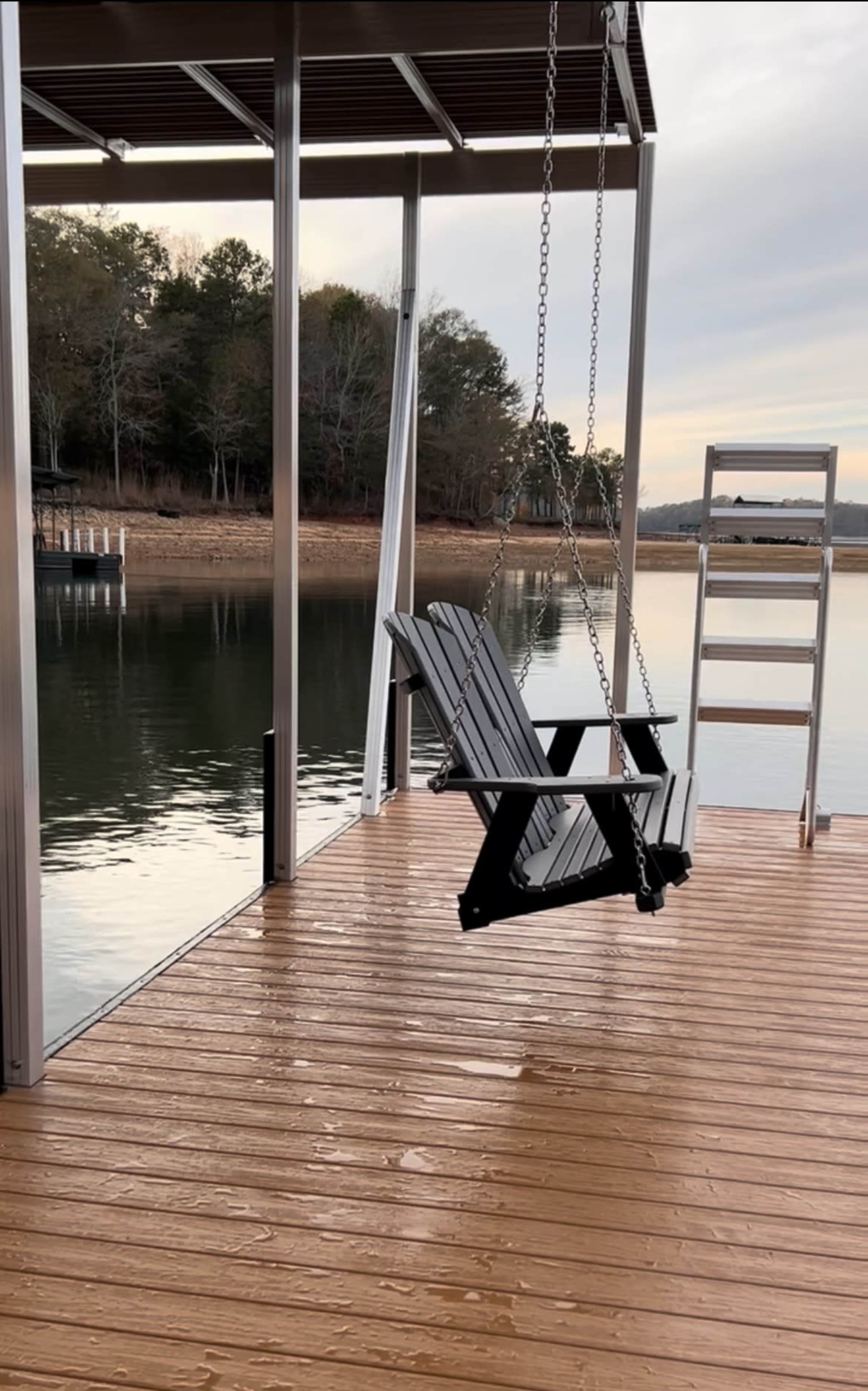 A black swing chair hangs from a dock overlooking a calm lake, with a metal ladder nearby and trees lining the distant shore.