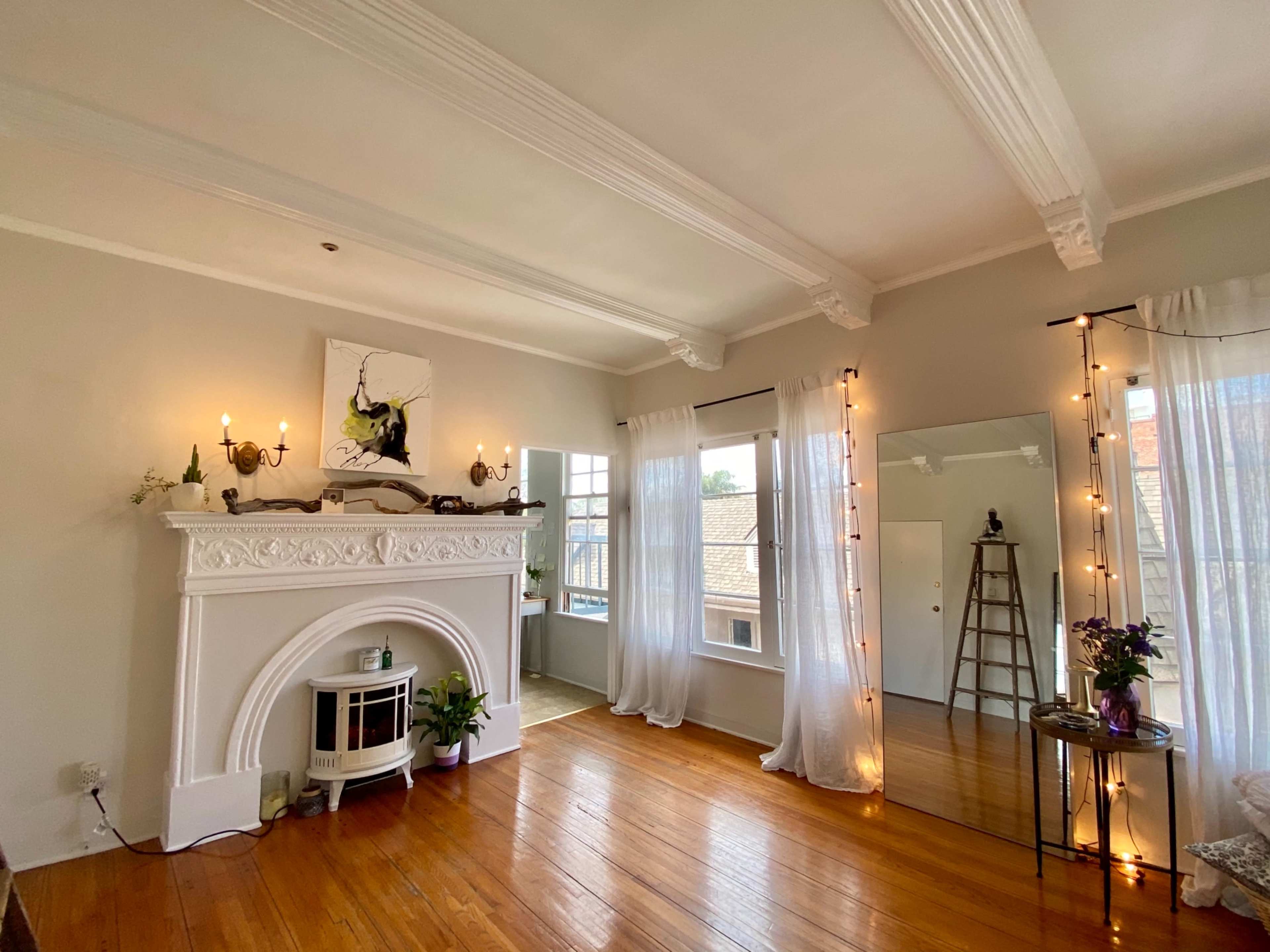 A light-filled room with a decorative fireplace, large windows, and a mirror reflecting a corner with a ladder and plants.