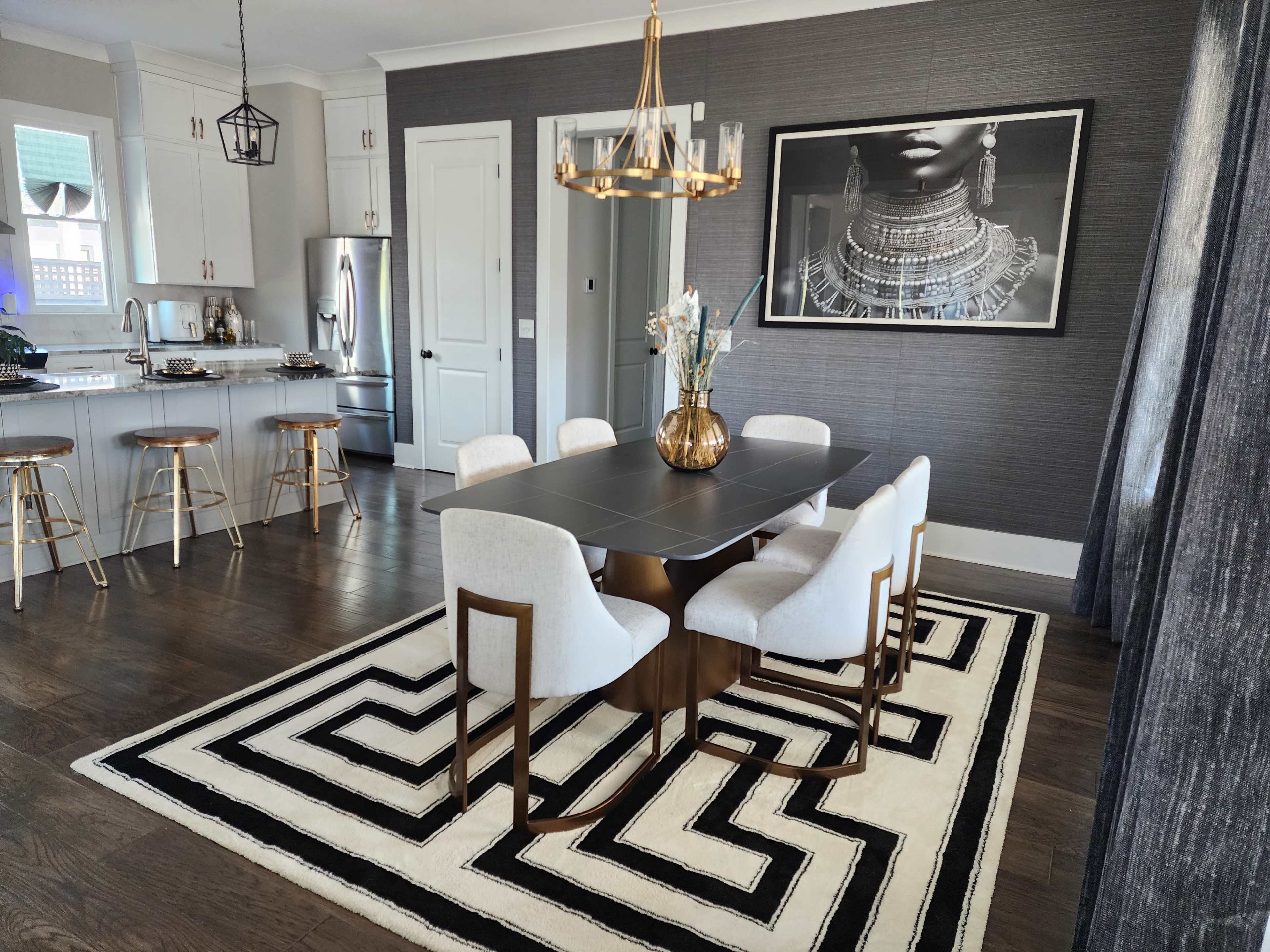 The image shows a modern dining area with a rectangular table surrounded by six chairs, a geometric rug, and a decorative chandelier overhead.