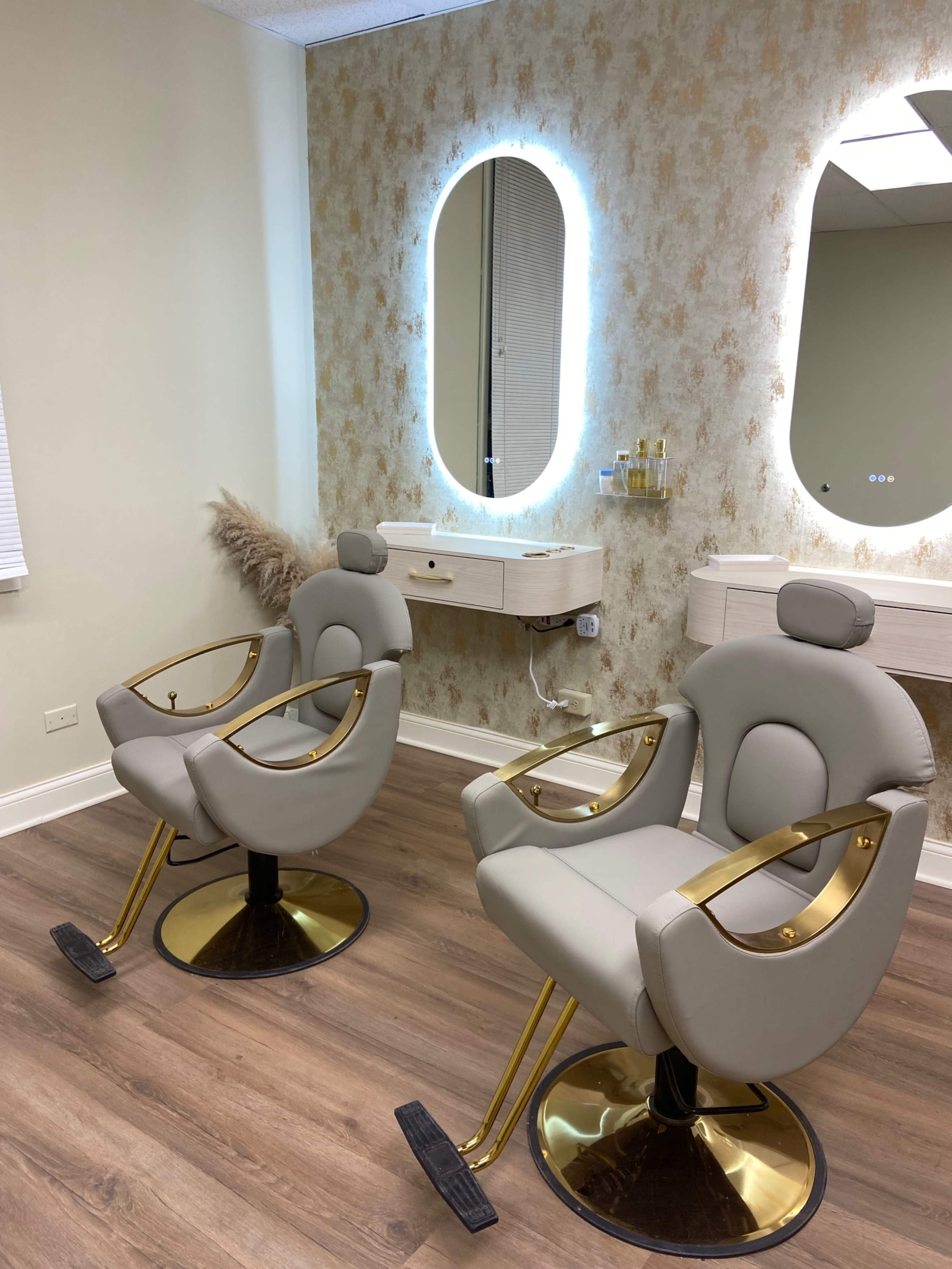 The image shows two modern salon chairs with gold accents placed in front of a mirror in a well-lit room with light-colored walls.