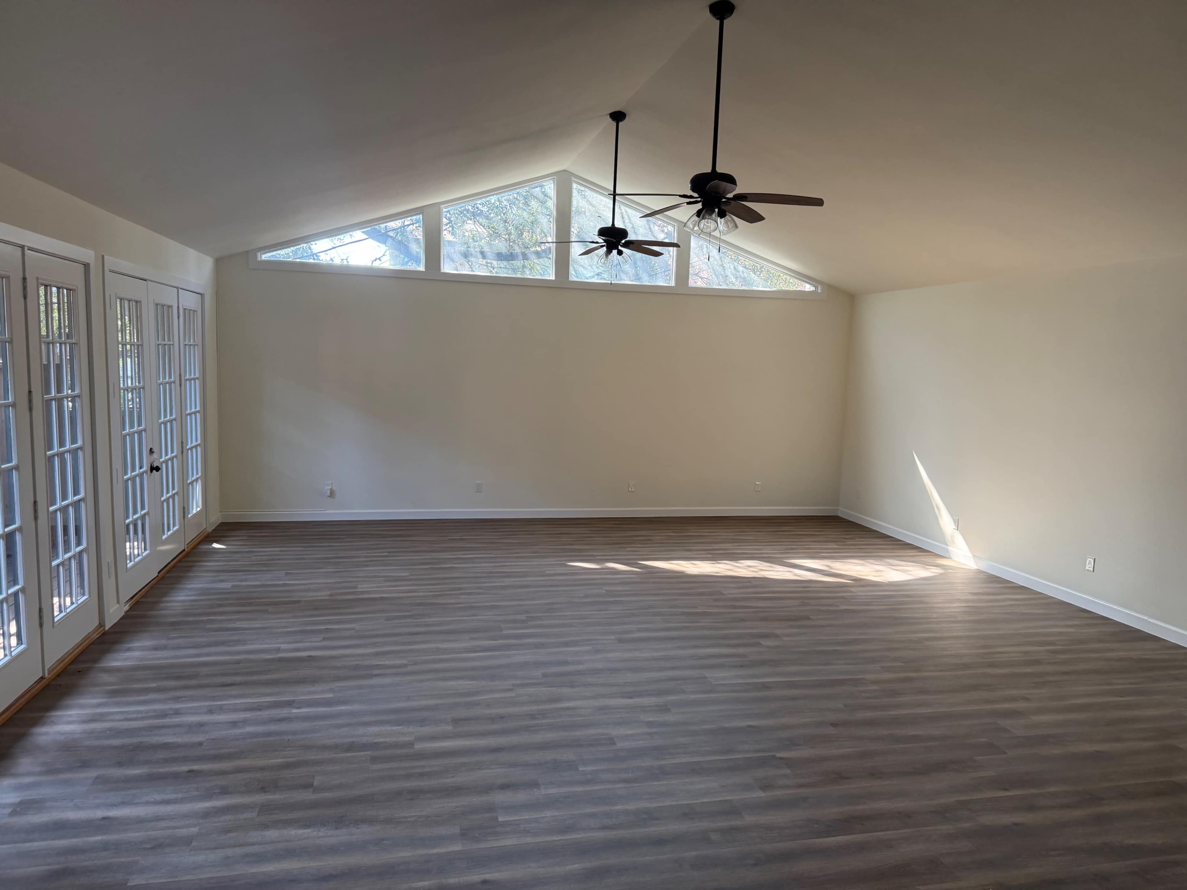 The image shows a spacious, empty room with a vaulted ceiling, two ceiling fans, large windows at one end, and French doors leading to an outdoor area.