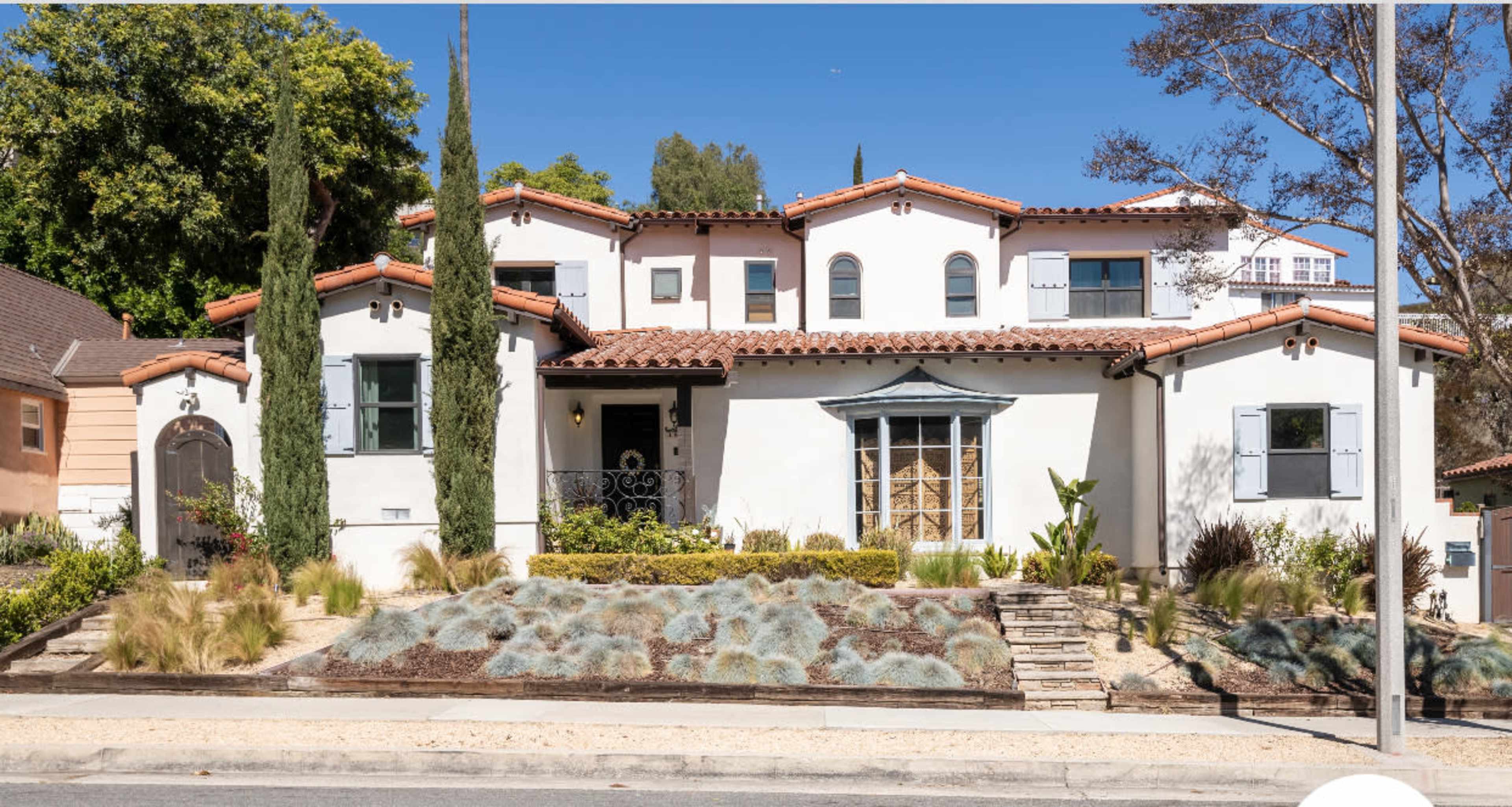 A two-story Mediterranean-style house with a terracotta roof, surrounded by neatly arranged landscaping and several tall cypress trees.