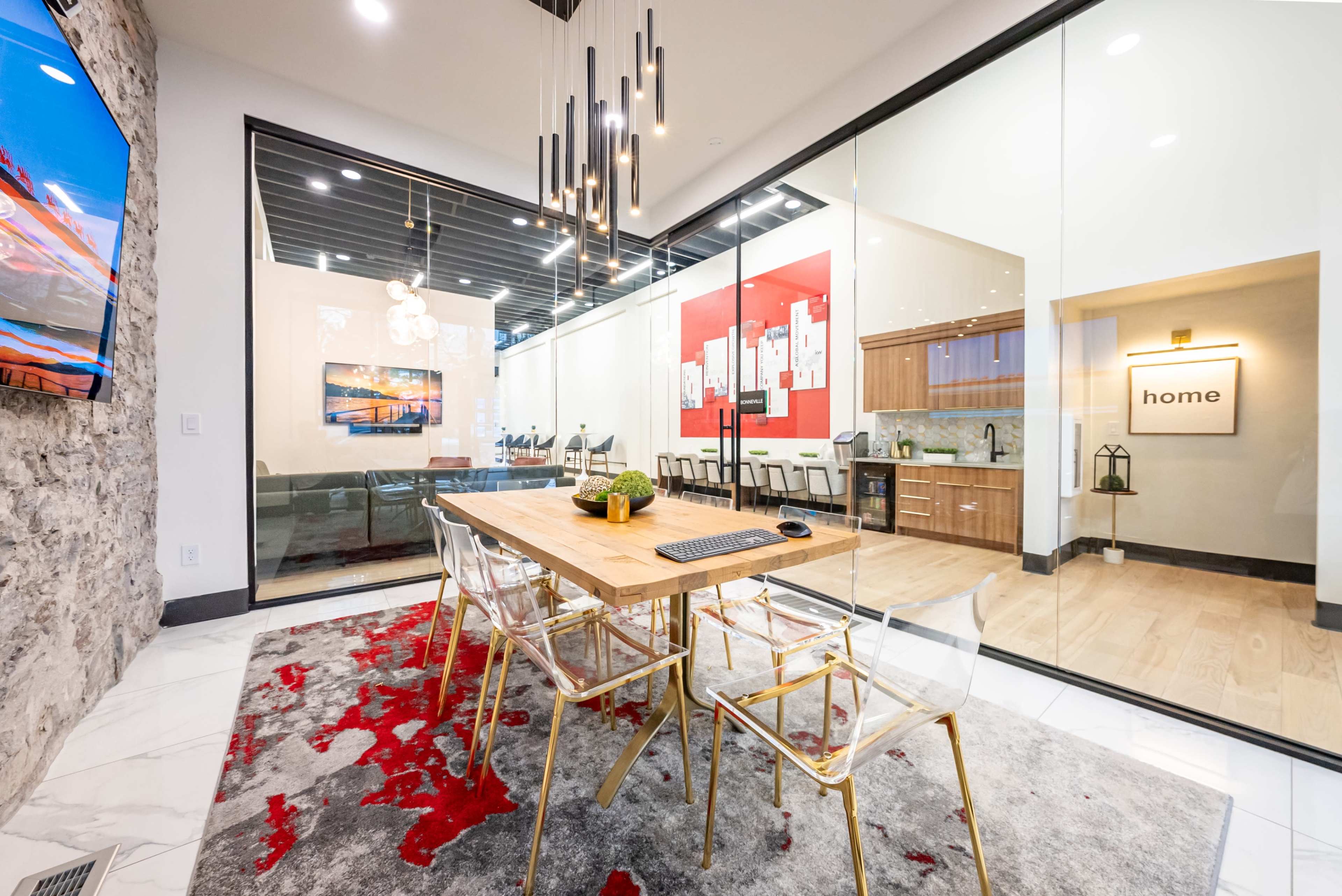 A modern conference room with a glass wall, featuring a wooden table surrounded by clear acrylic chairs, a large screen on one wall, and a kitchen area visible in the background.