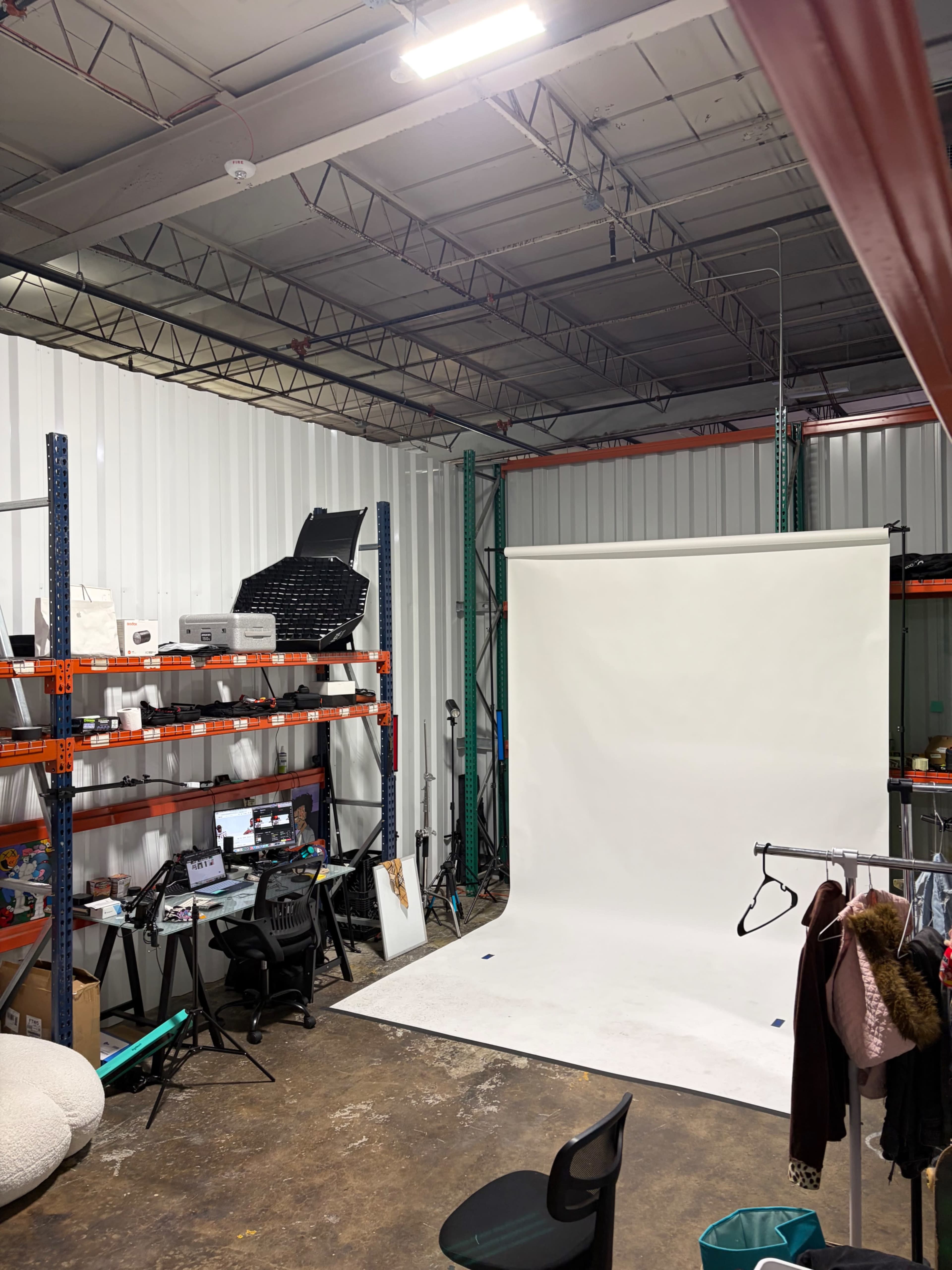 The image shows a photography studio with a plain white backdrop, shelves filled with equipment, and a workspace featuring computer monitors and a chair.