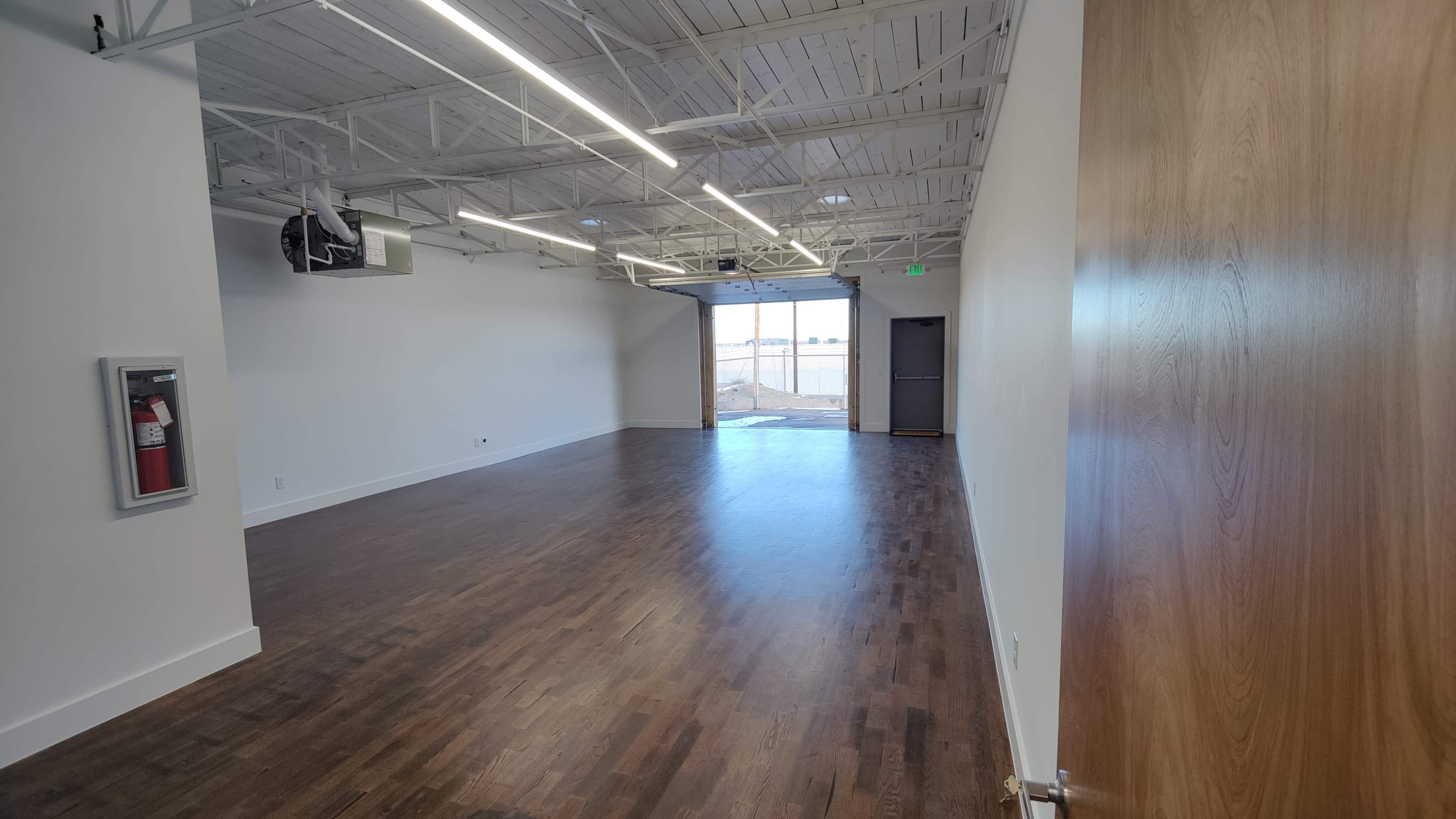 The image shows an empty interior room with hardwood flooring and large windows at the far end, allowing natural light to enter.