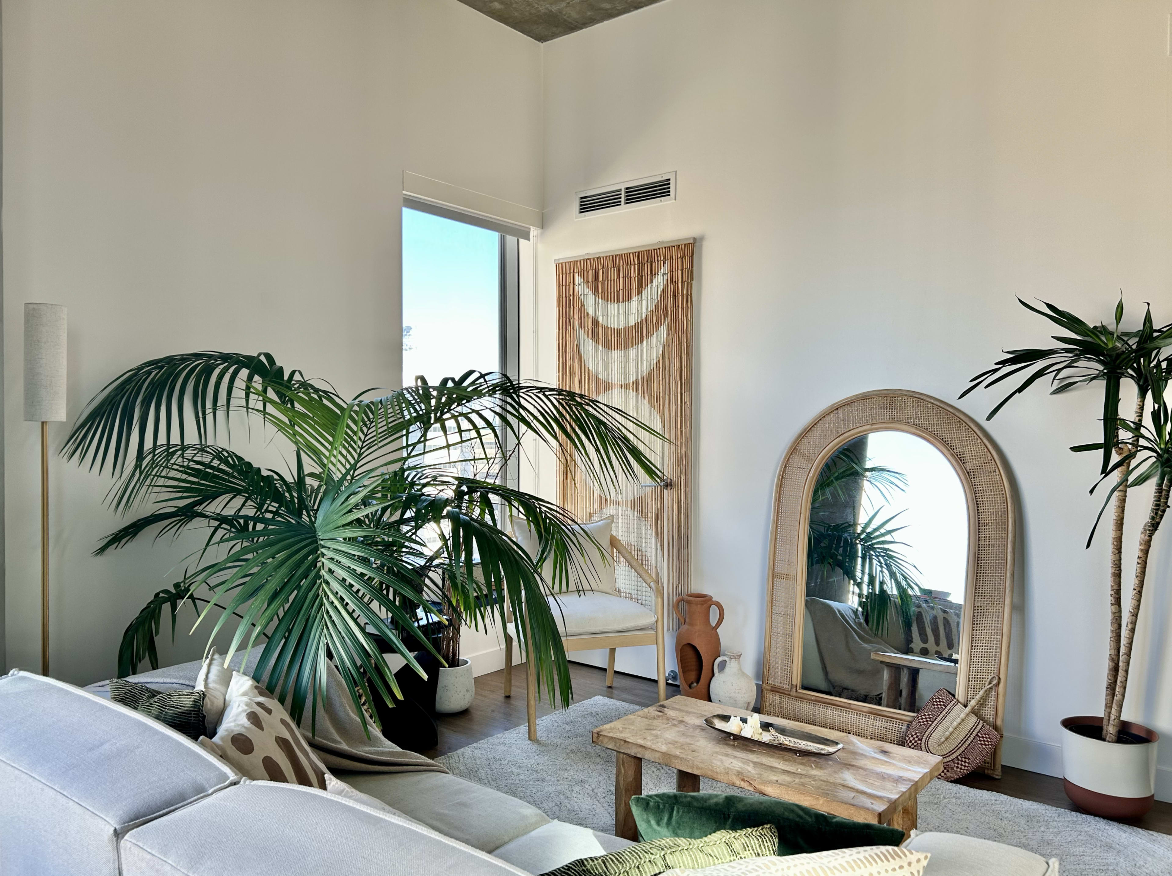 The image shows a modern living room with a large mirror, plants, a wooden coffee table, and a comfortable sofa.