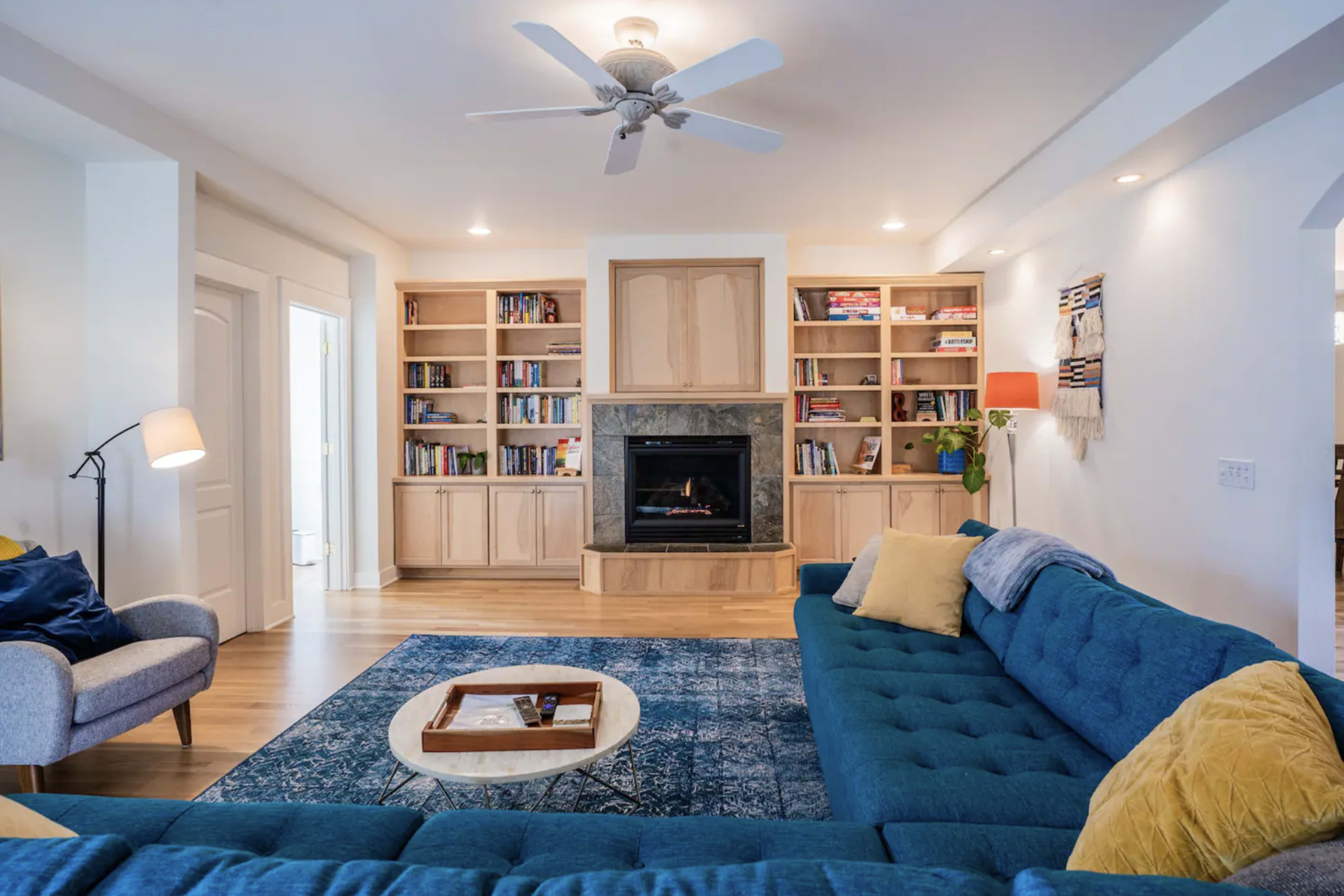 A living room features a blue sectional sofa, a fireplace surrounded by built-in bookshelves, and a decorative coffee table on a patterned rug.