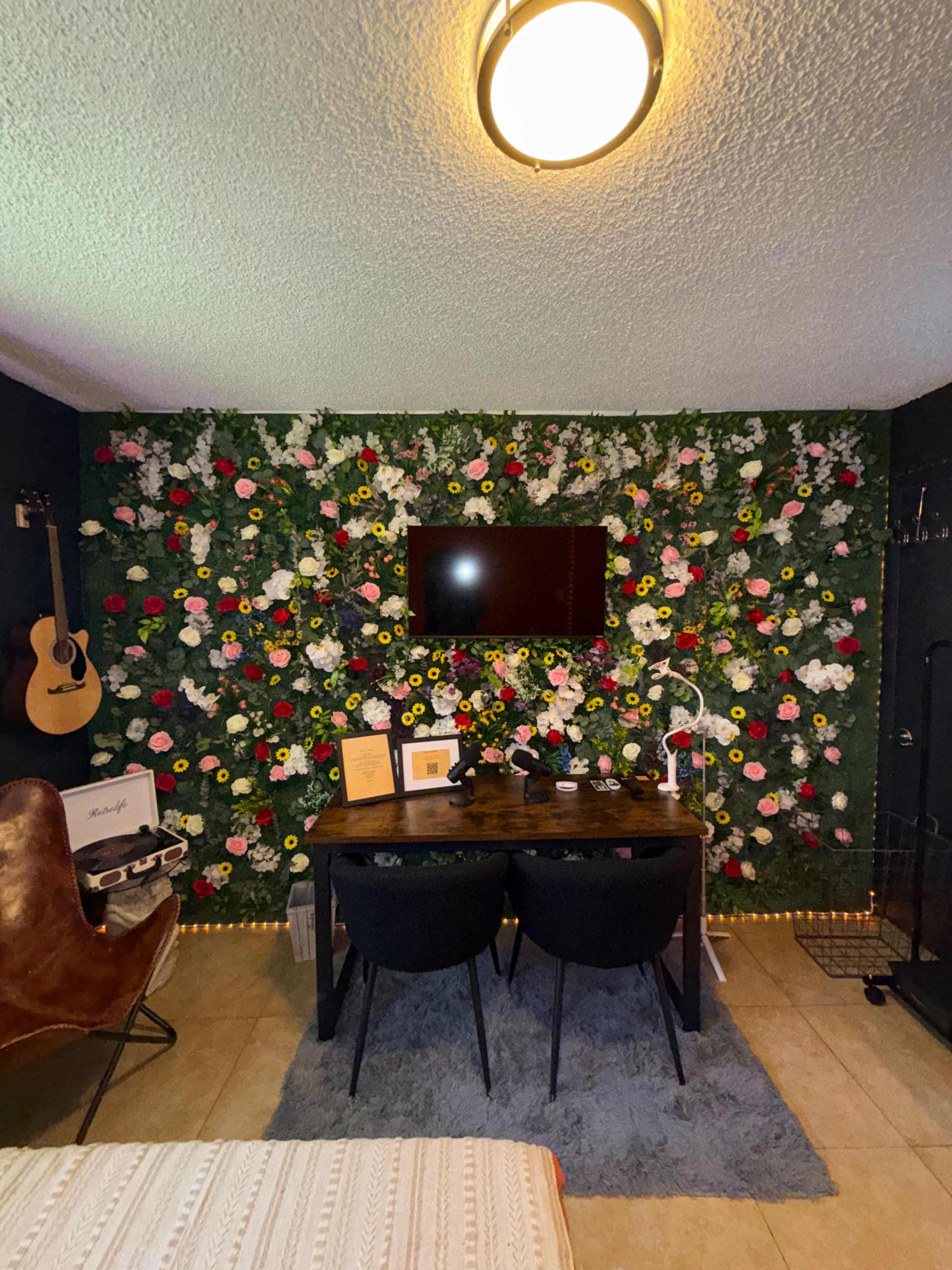 The image shows a home workspace with a floral wall, a wooden desk with two black chairs, and a television mounted above the desk.