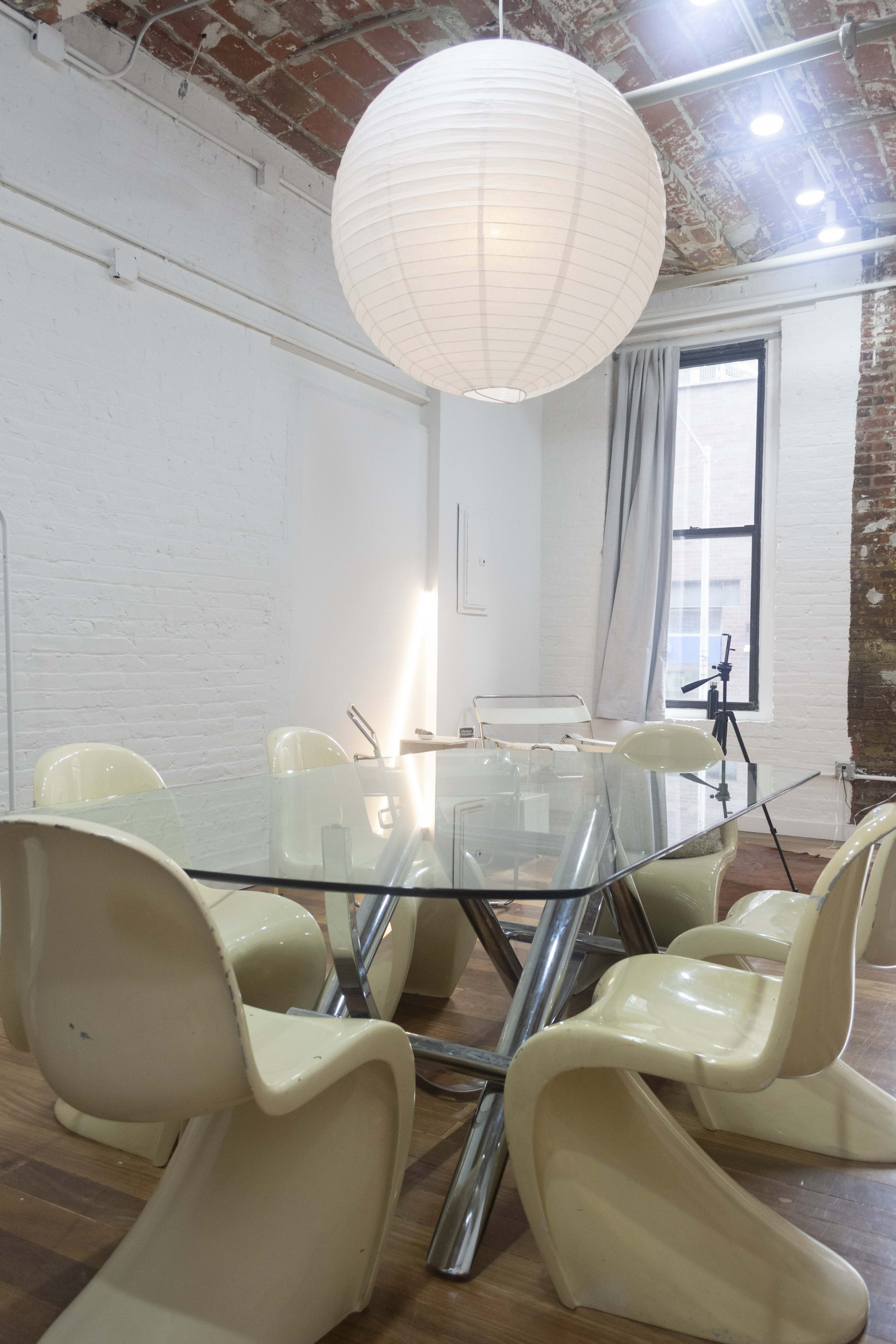 A glass dining table with six yellow chairs is illuminated by a large round pendant light in a room with exposed brick walls.
