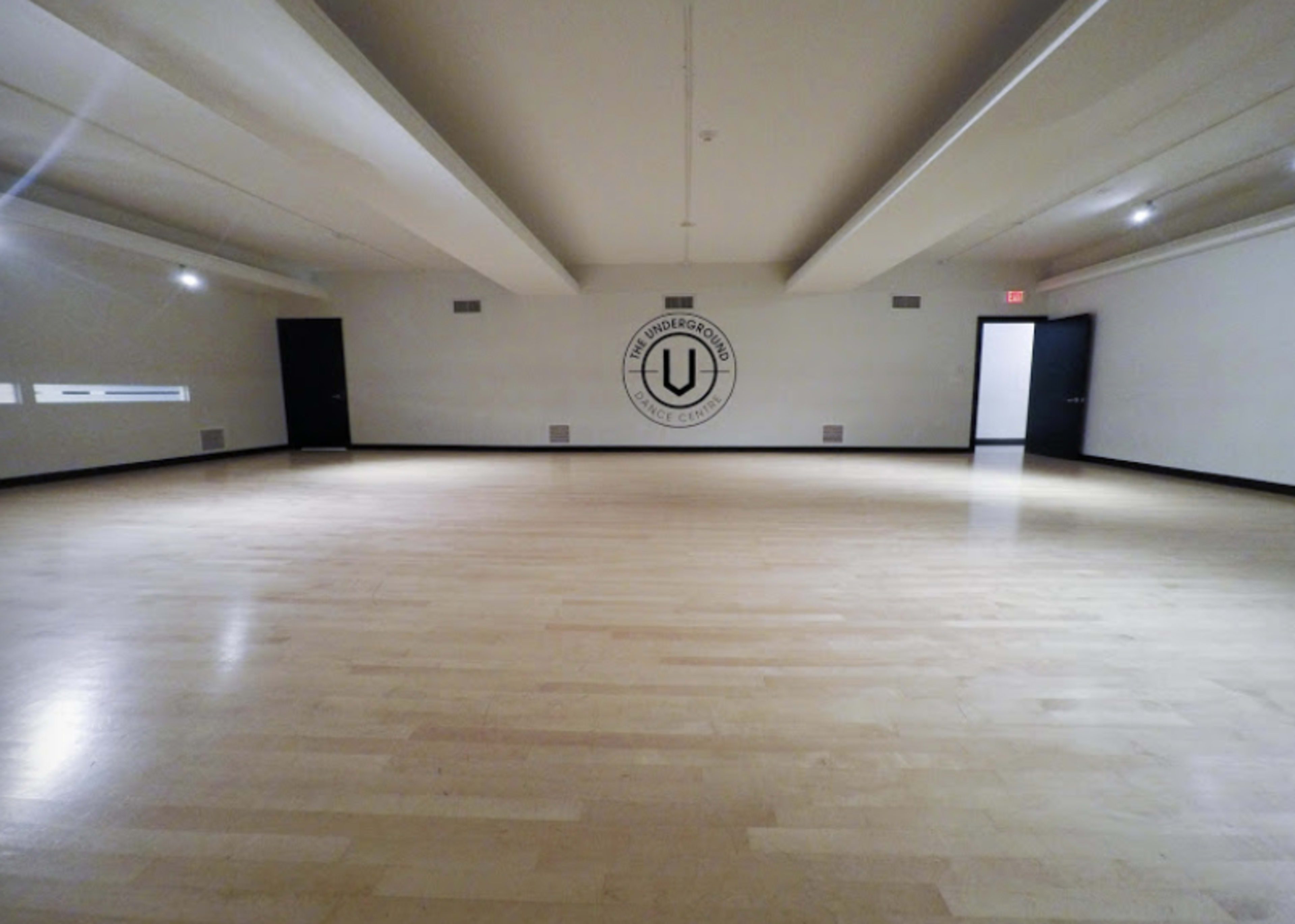 A large, empty dance studio features wooden flooring and minimal lighting, with a logo on the wall and two doors on opposite sides.
