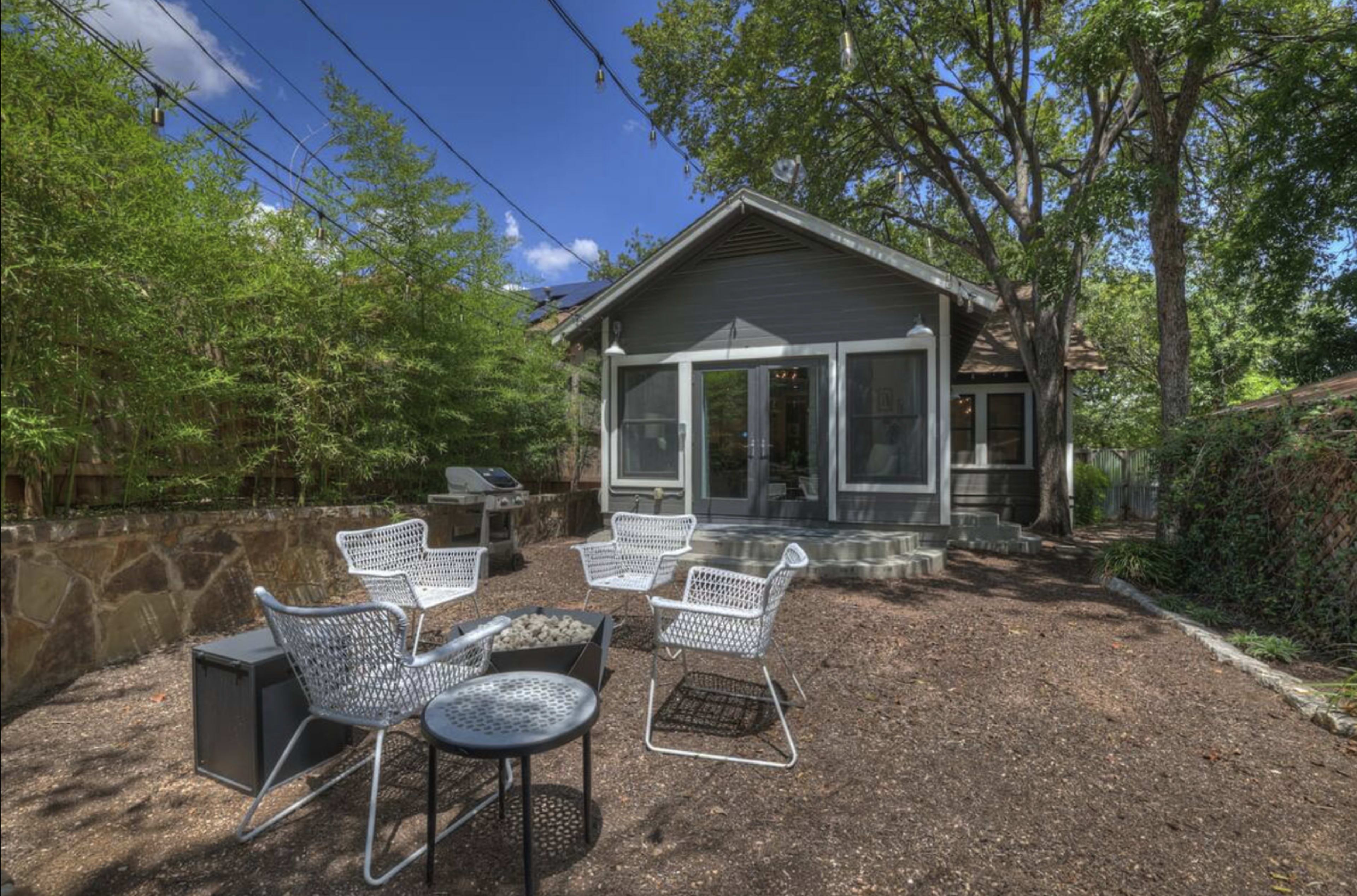 A backyard features a modern home with a patio area surrounded by several outdoor chairs and a small table, complemented by greenery and a barbecue grill.