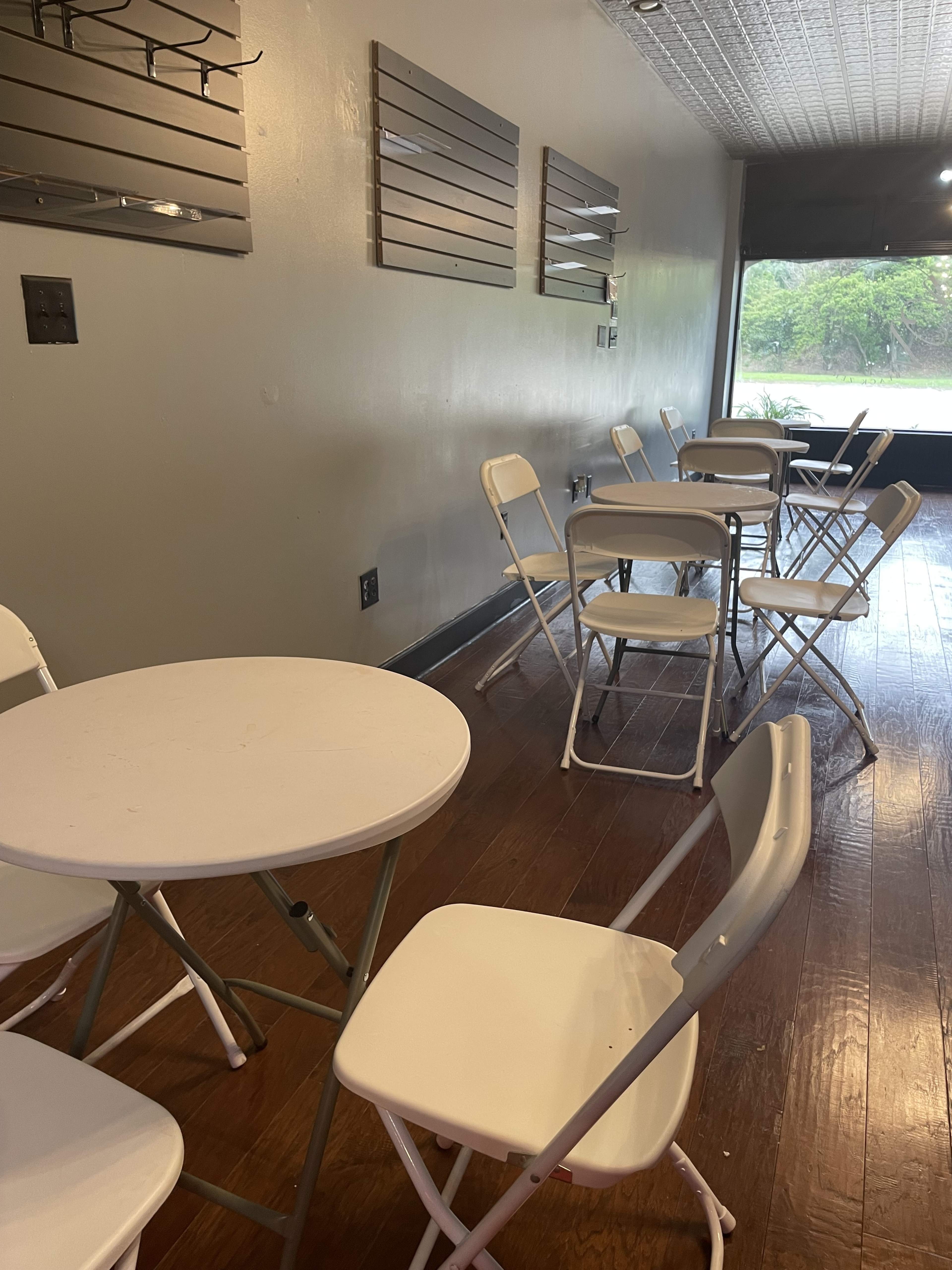 The image shows a sparsely furnished room with several white folding tables and chairs arranged around a wooden floor.