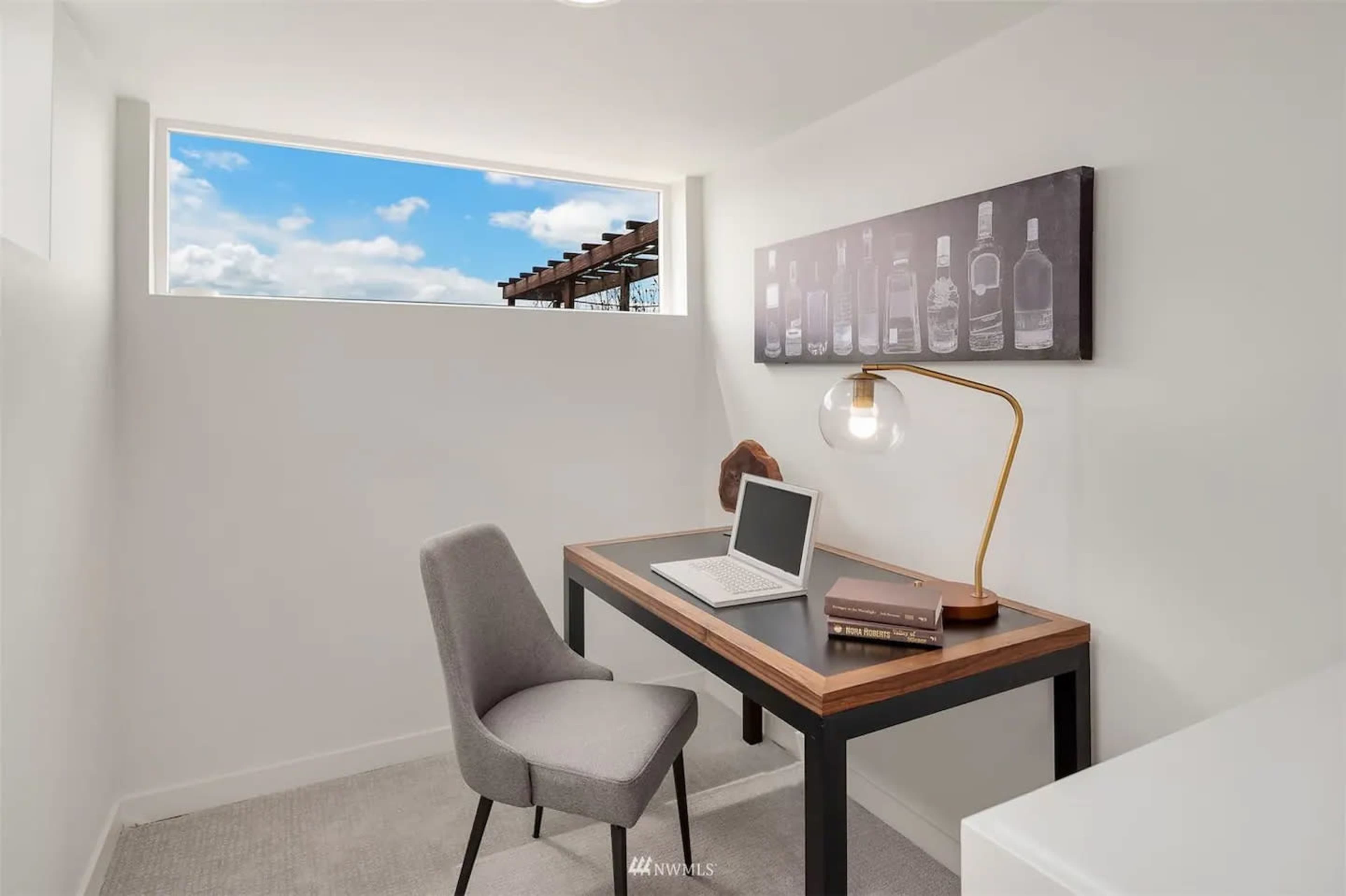 A small, well-lit workspace featuring a desk with a laptop, a decorative lamp, and a bottle-themed artwork on the wall.