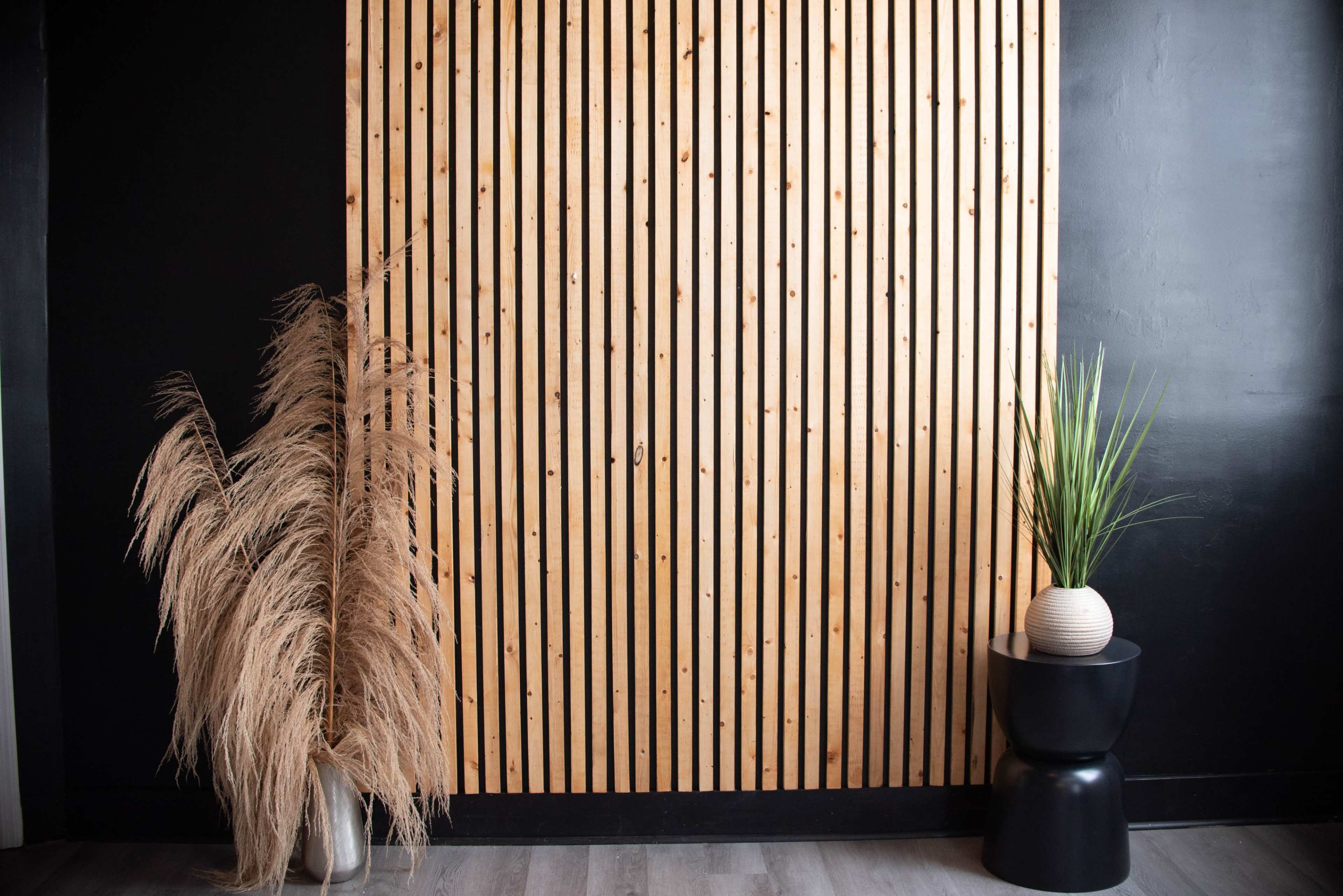 A wooden slatted wall, a tall pampas grass arrangement, and a potted plant on a round black table.