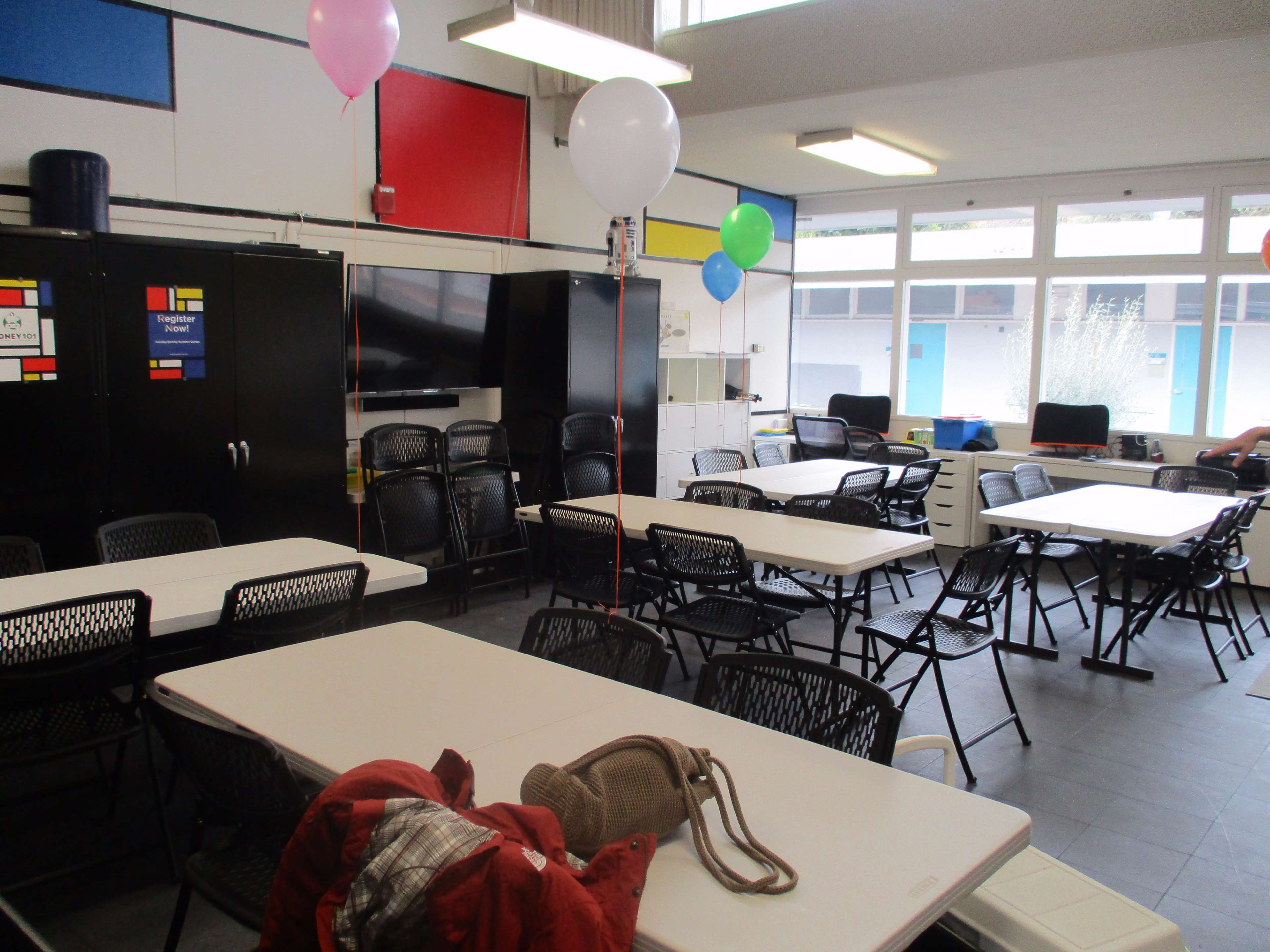 A room with several tables and chairs, decorated with balloons, and featuring a television and storage units.