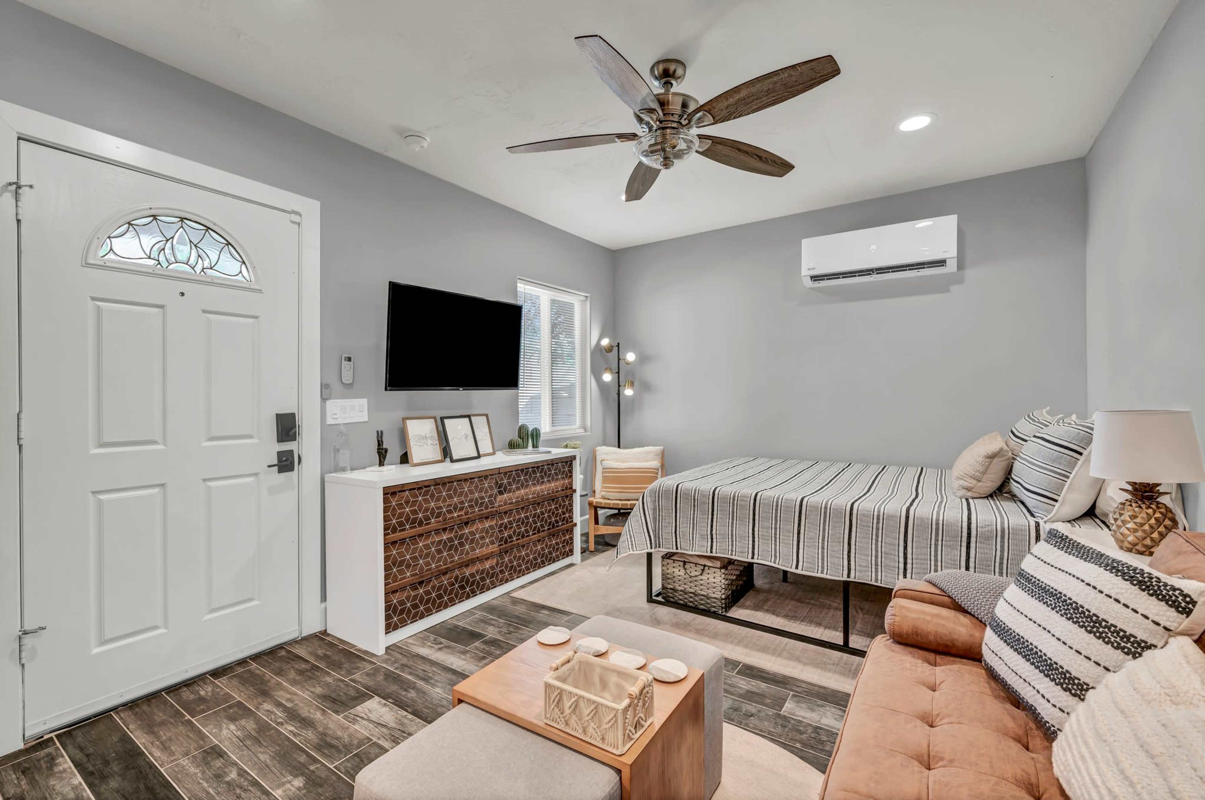 A small living space featuring a bed, a sofa, a television mounted on the wall, and a decorative dresser, with a front door visible.
