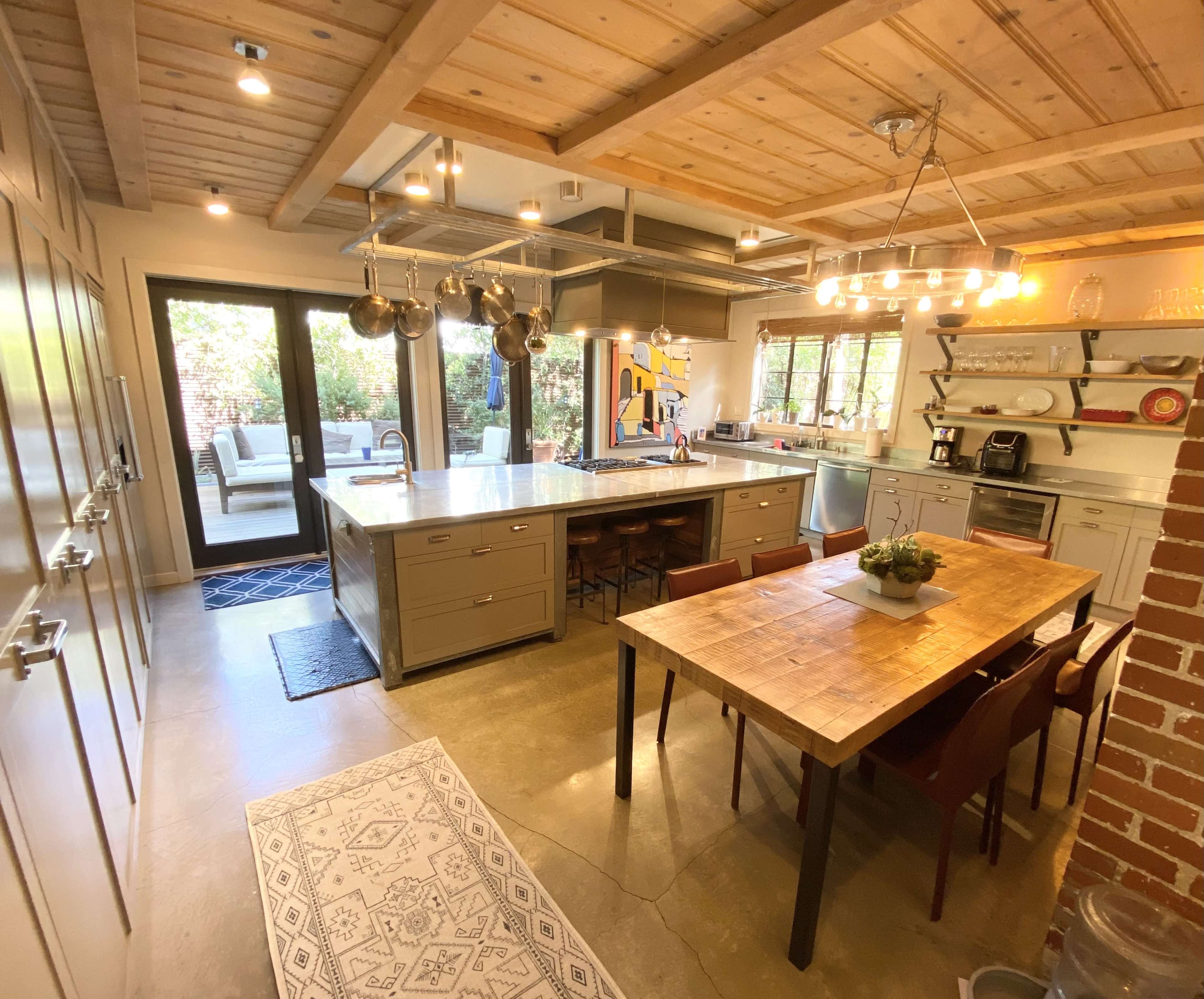 A modern kitchen features a large island, wooden dining table, and natural light from sliding glass doors opening to an outdoor space.