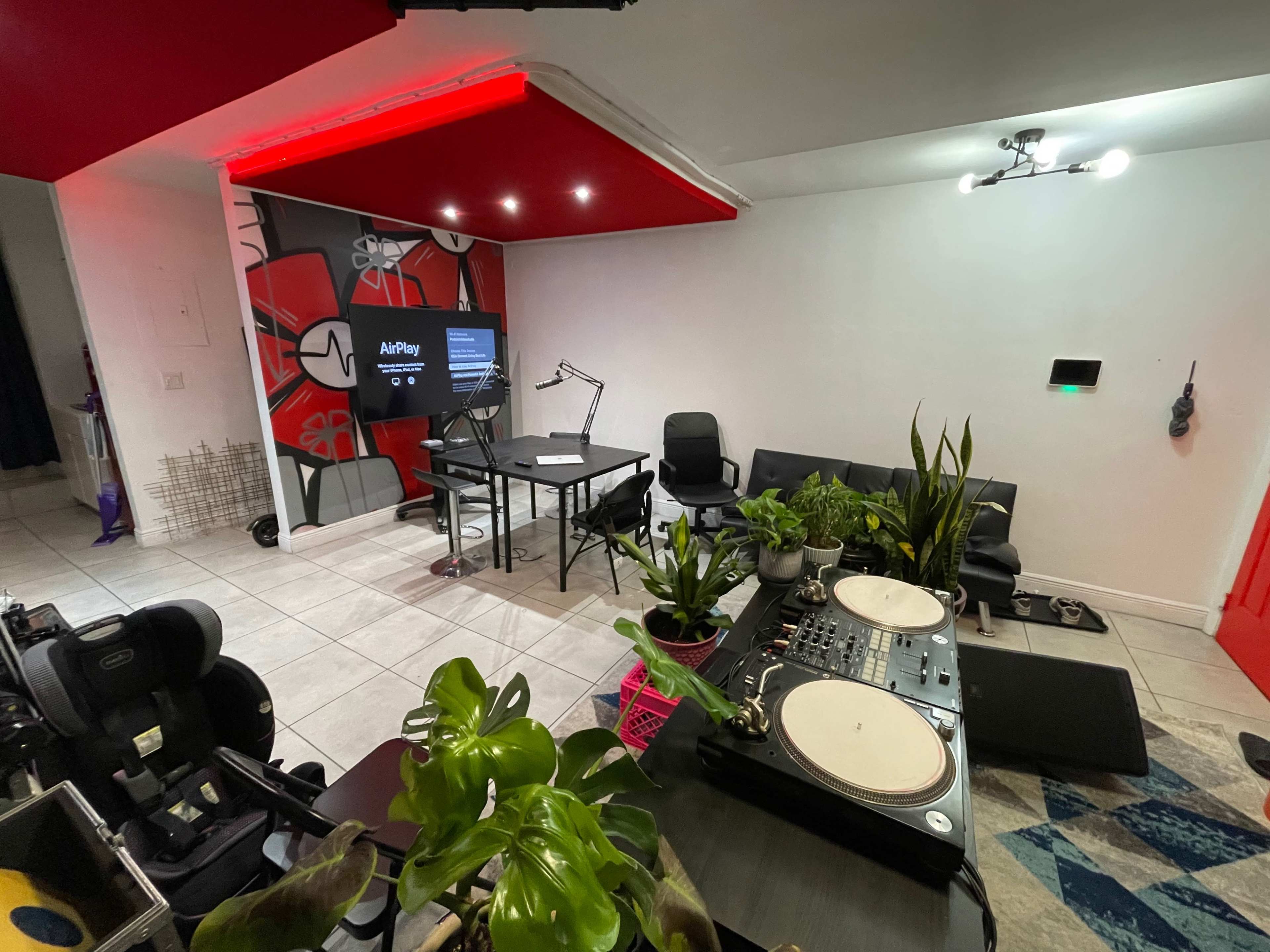 A stylish room set up with a DJ table, a large screen displaying "AirPlay," several plants, and contemporary seating arrangements.